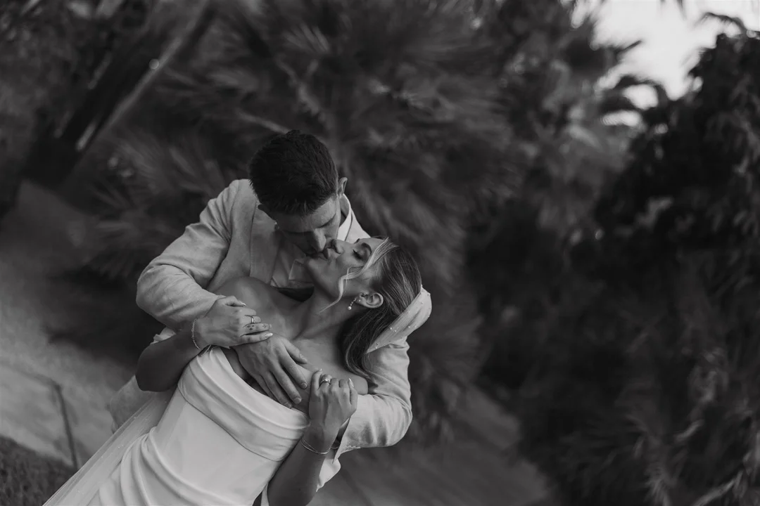 A black and white photo of a couple sharing a kiss, with the man holding the woman in a dance pose outdoors near trees.