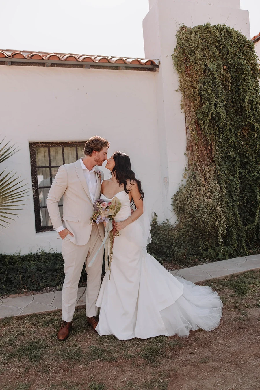 A bride and groom in wedding attire standing close and touching noses outdoors next to a white building with a window and greenery.
