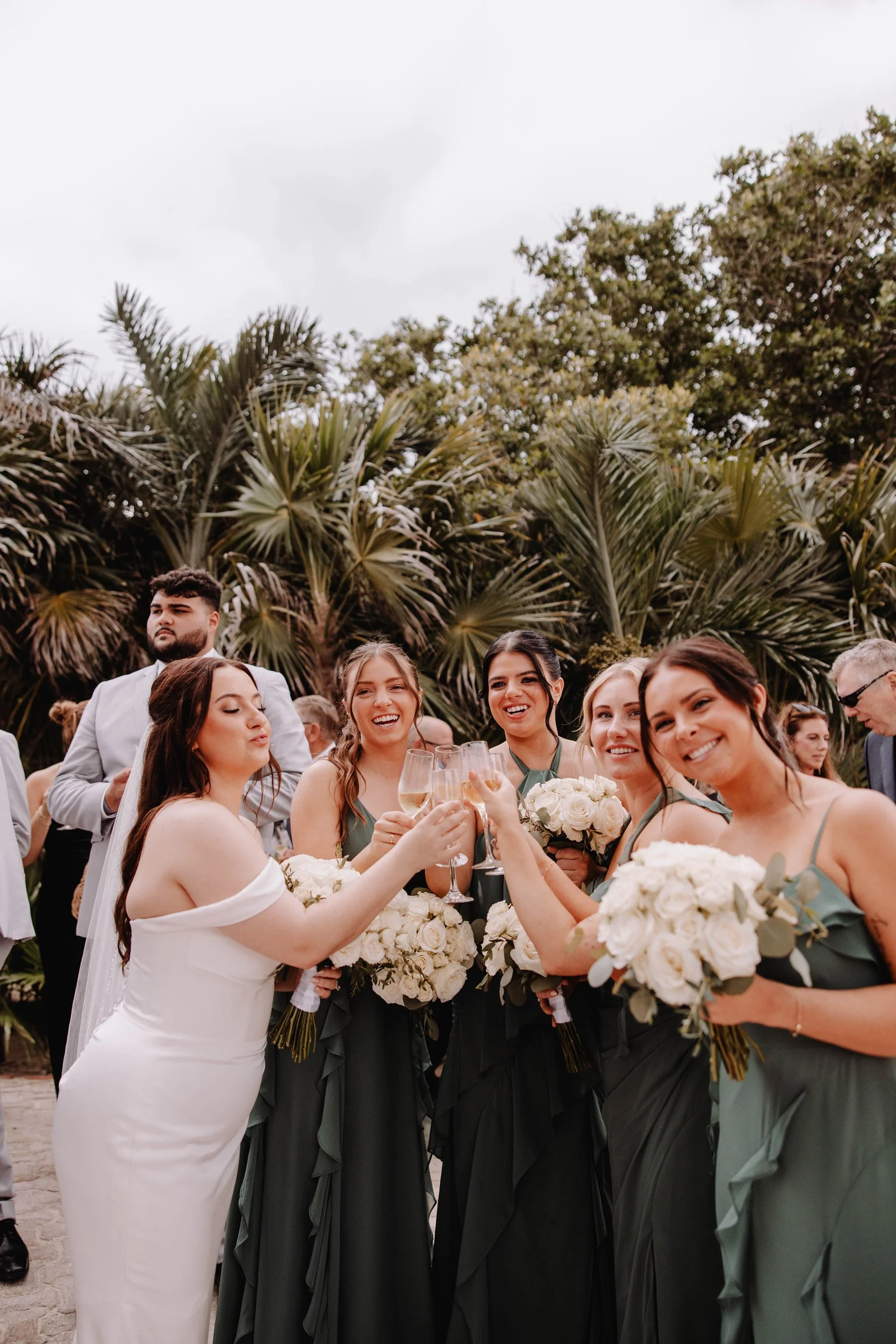 Group of women at a wedding celebration, holding bouquets and champagne glasses, toasting and smiling outdoors with palm trees in the background.