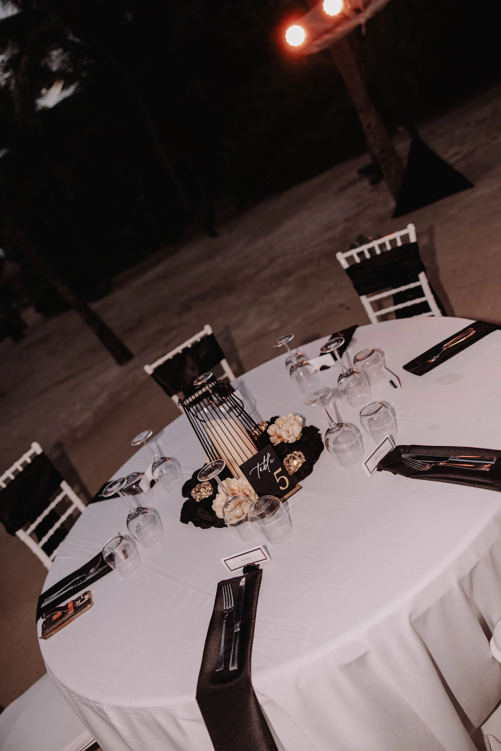 A round table set for a formal event outdoors at night, with a white tablecloth, black napkins, glassware, and decorative centerpieces including flowers and a table number card.