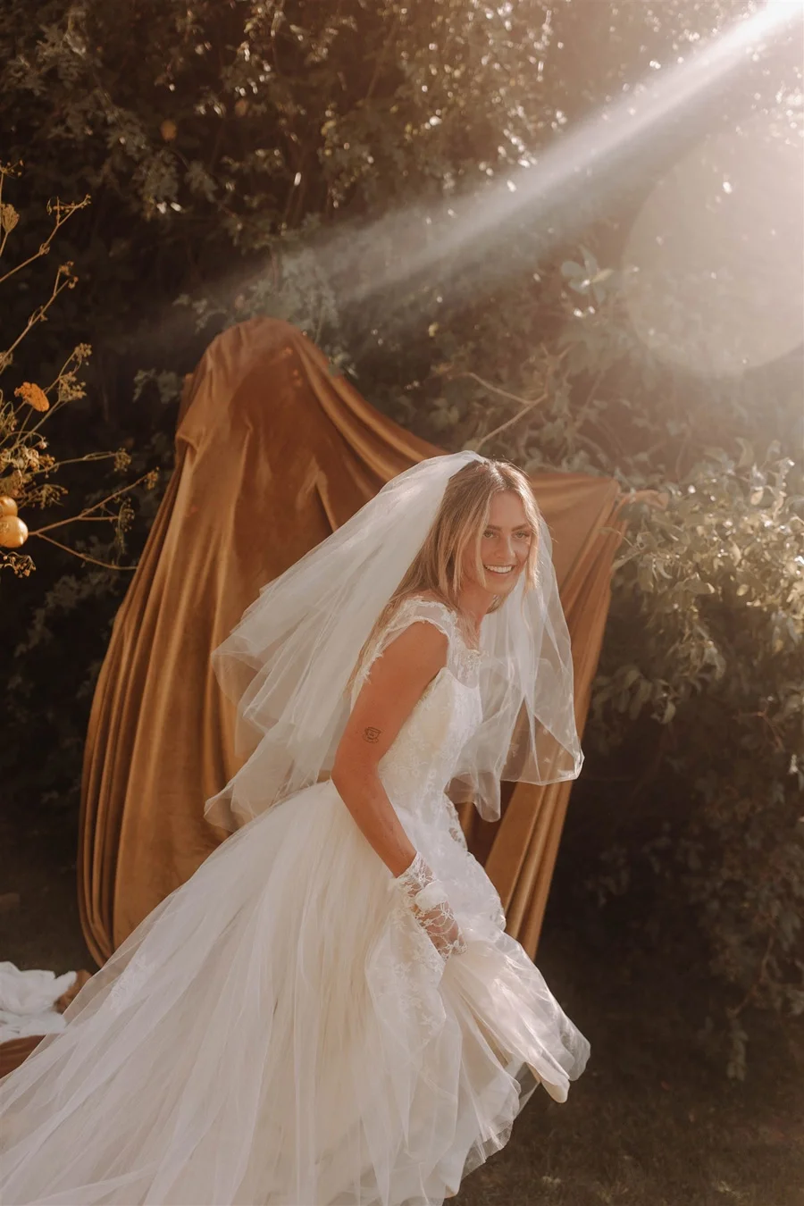A woman in a wedding dress with a veil smiling outdoors in front of a large brown fabric backdrop, with sunlight streaming from the right side.