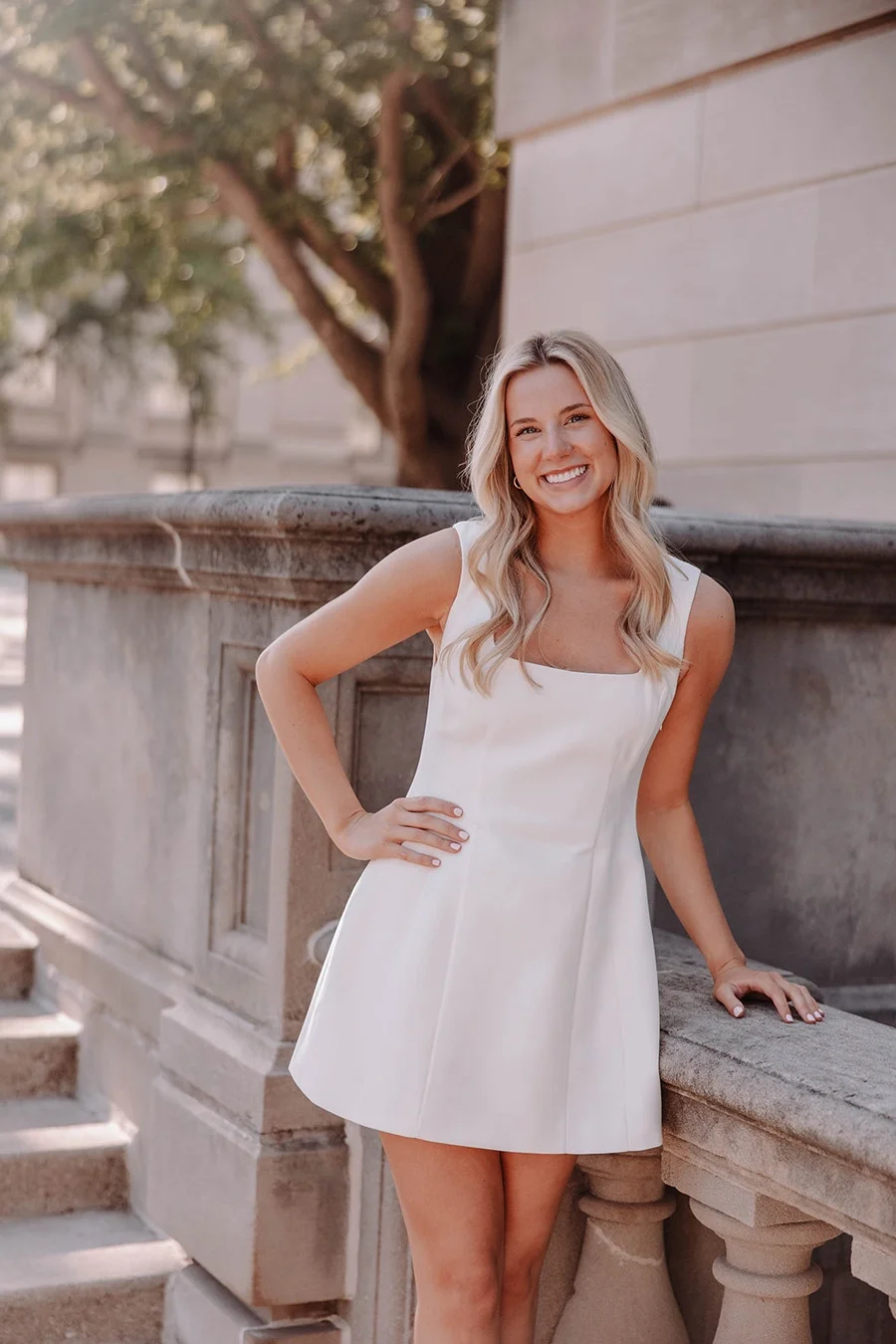 A young woman with long blonde hair, smiling in a white sleeveless dress, standing outdoors next to a stone railing with steps, in front of a building and trees.