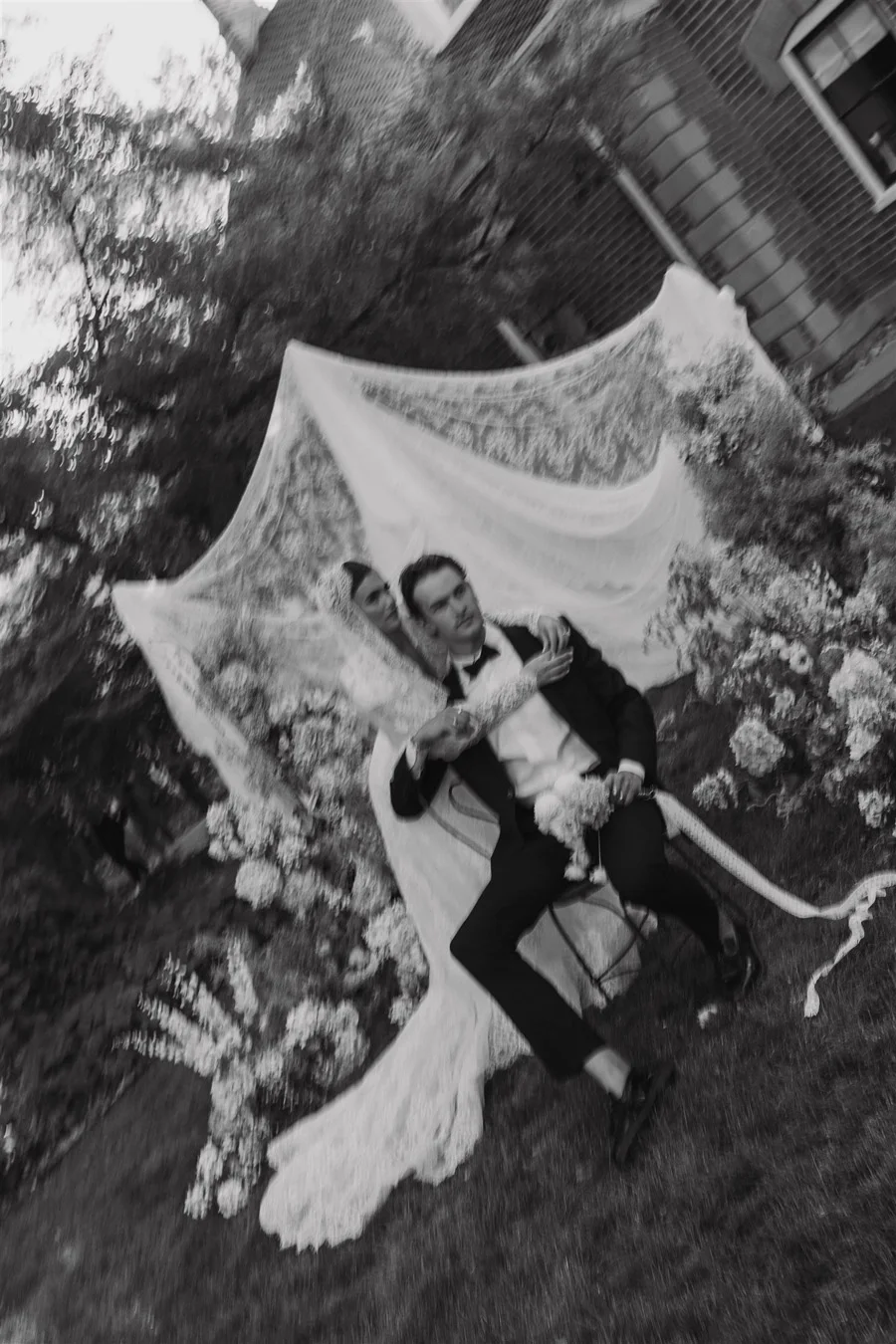 A person in formal attire, sitting on a chair, is posing in front of a decorative backdrop with a large portrait of a woman. The scene appears to be an outdoor event with plants and a building in the background, and it is captured in black and white.