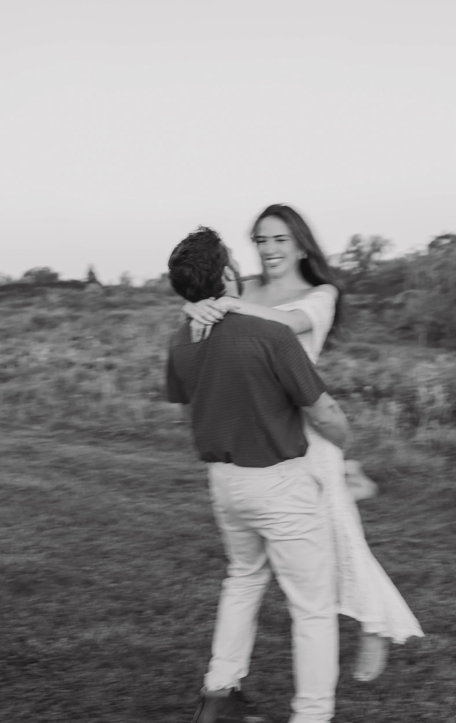 A couple is outdoors, with the man lifting the woman off the ground. They are smiling and embracing, with a blurred natural landscape in the background, captured in black and white.