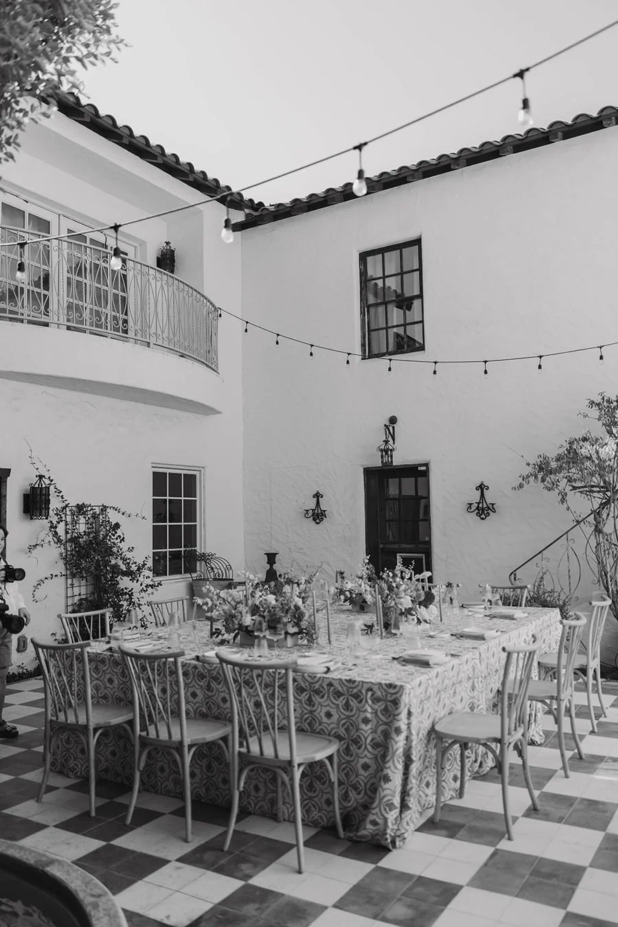 An outdoor patio with a long dining table covered with a patterned tablecloth, decorated with flower arrangements, and surrounded by chairs. Above, string lights are hung across the area. The building features a balcony with a wrought iron railing, windows, and wall-mounted lanterns. The scene appears to be set for a gathering or celebration.
