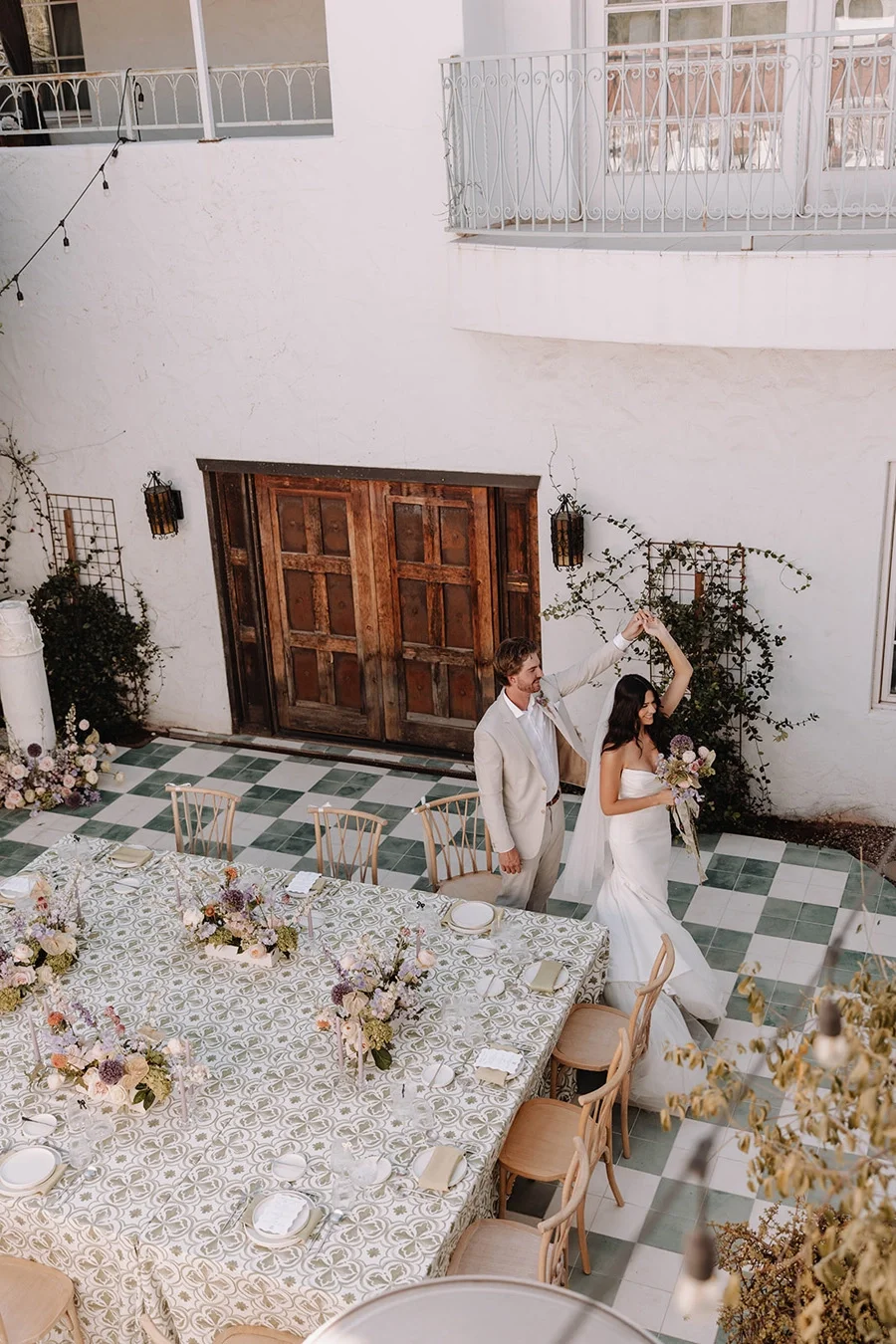 A bride and groom are dancing and holding hands inside a decorated dining area, with tables set for a wedding celebration.