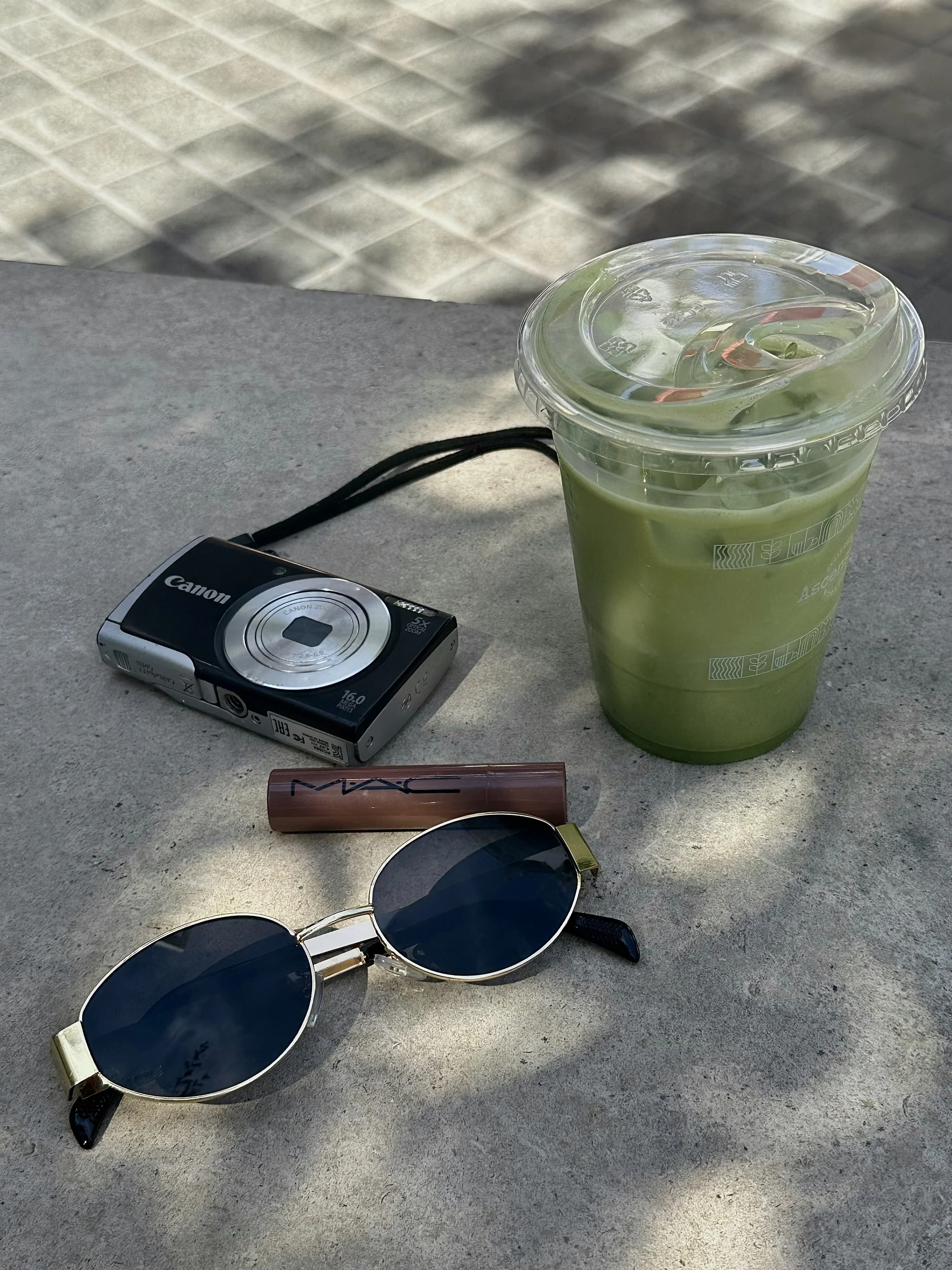 A plastic cup of green iced matcha latte on a table next to sunglasses, a vintage digital camera, and a tube of MAC lipstick in an outdoor setting with a tiled pavement in the background.
