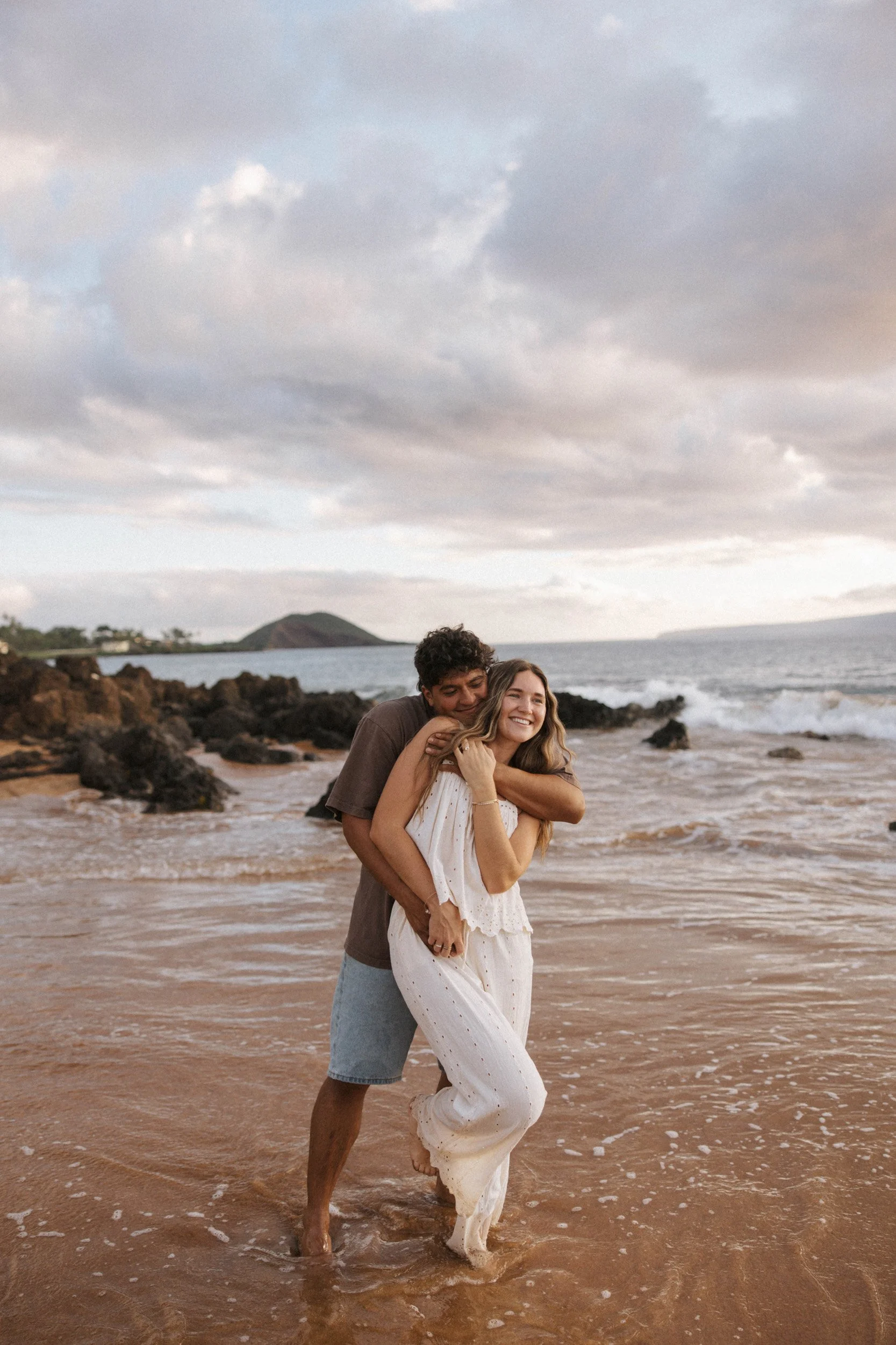 A young couple on the beach, smiling and embracing each other, with the ocean, rocks, and a cloudy sky in the background.