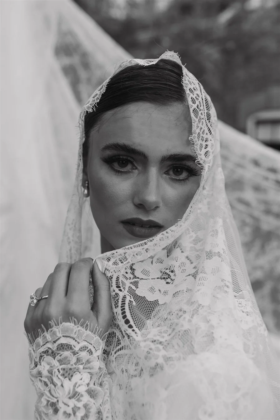 A woman with dark hair, wearing lace clothing and a lace head covering, looking directly at the camera while holding the edge of her lace head covering near her face.