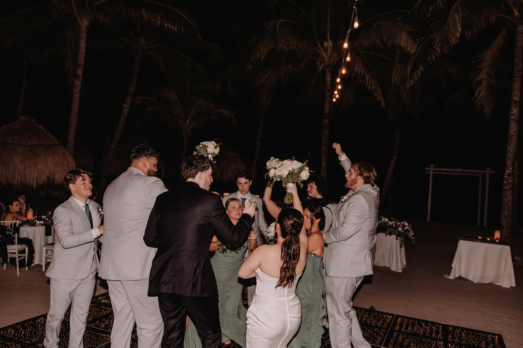 Group of people celebrating at a wedding reception, some holding bouquets, outdoors at night with string lights and palm trees.