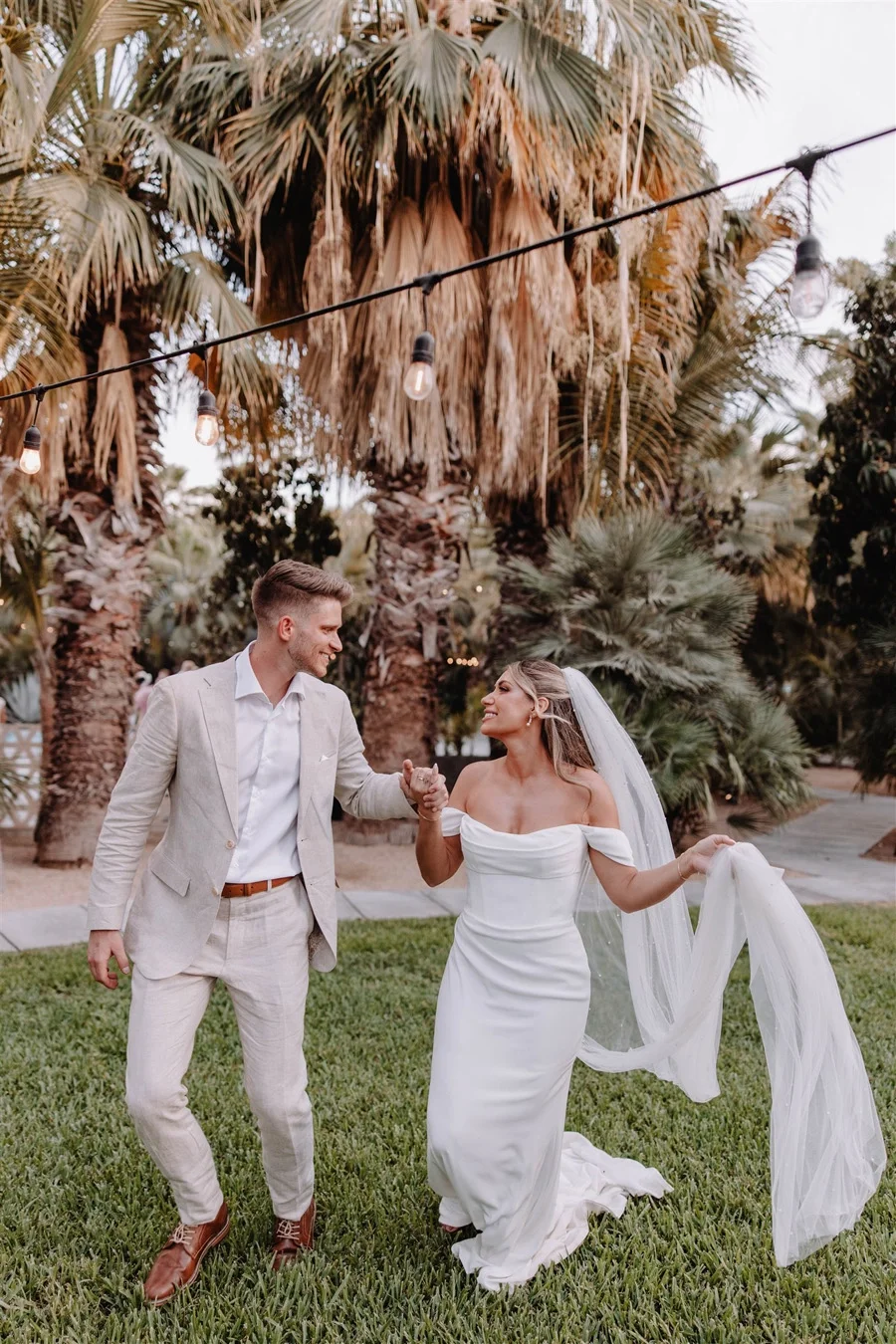 A bride and groom dancing outdoors on a grassy area with palm trees and string lights overhead, celebrating their wedding.