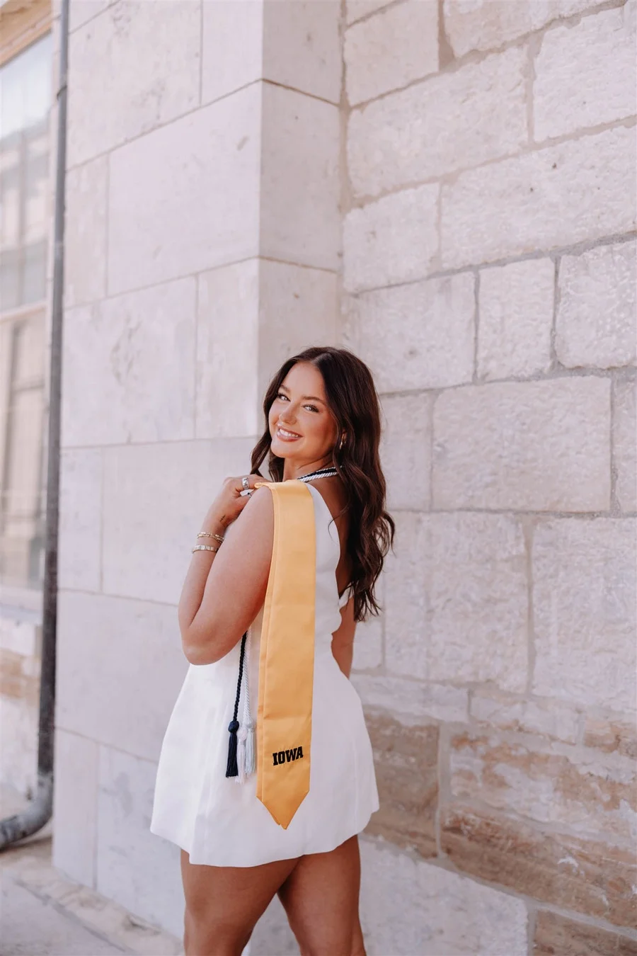 A woman in a white dress with an Iowa graduation stole, smiling and looking over her shoulder, stands against a stone building.