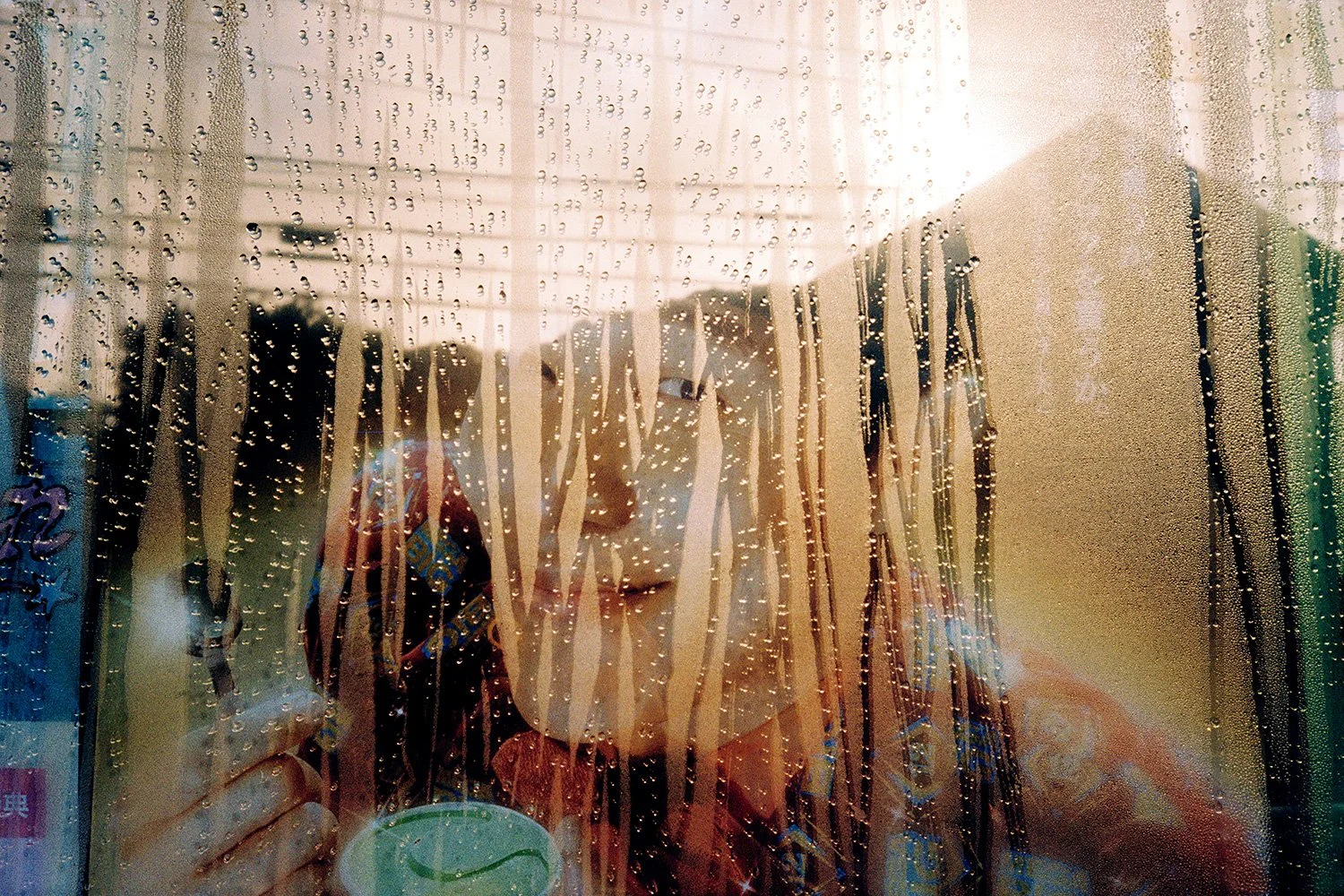 A person with glasses is seen through a rain-covered glass door with ornaments hanging, causing the person's reflection and the background to appear distorted and blurry.
