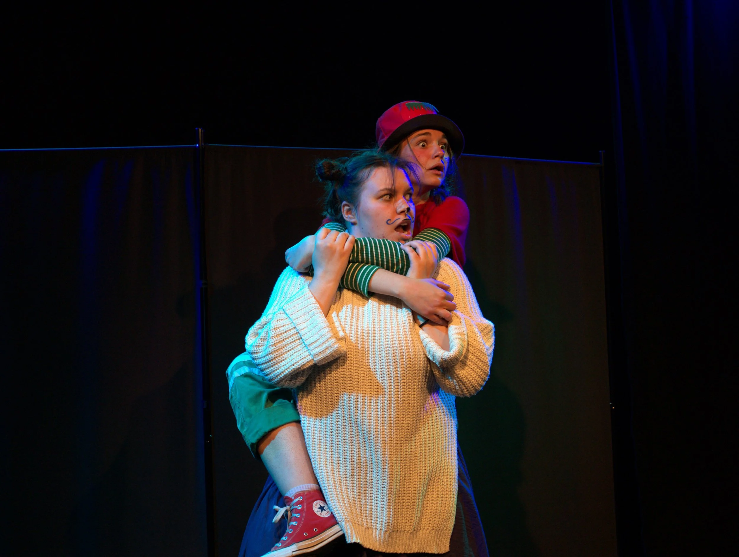 Deux jeunes femmes expriment la surprise ou l'inquiétude, l'une portant l'autre dans ses bras, lors d'une performance sur scène avec un fond noir.