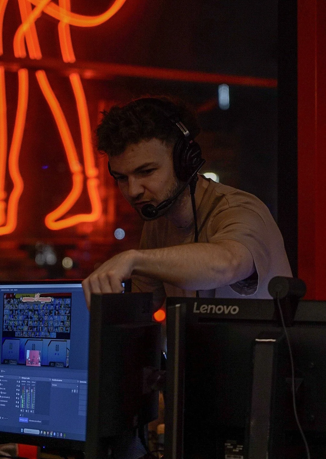 Un homme portant un casque avec micro, utilisant un ordinateur dans un environnement sombre avec un néon orange en arrière-plan.