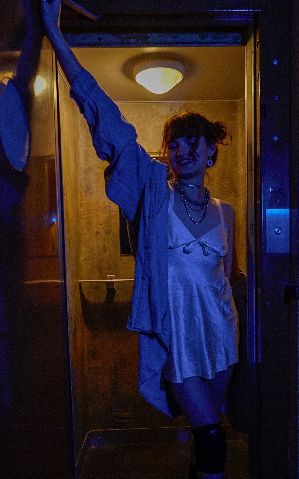 Une femme souriante portant une robe blanche avec un haut en coton blanc et un collier multi-rangs, se trouve dans un ascenseur sombre éclairé par une lumière jaune.