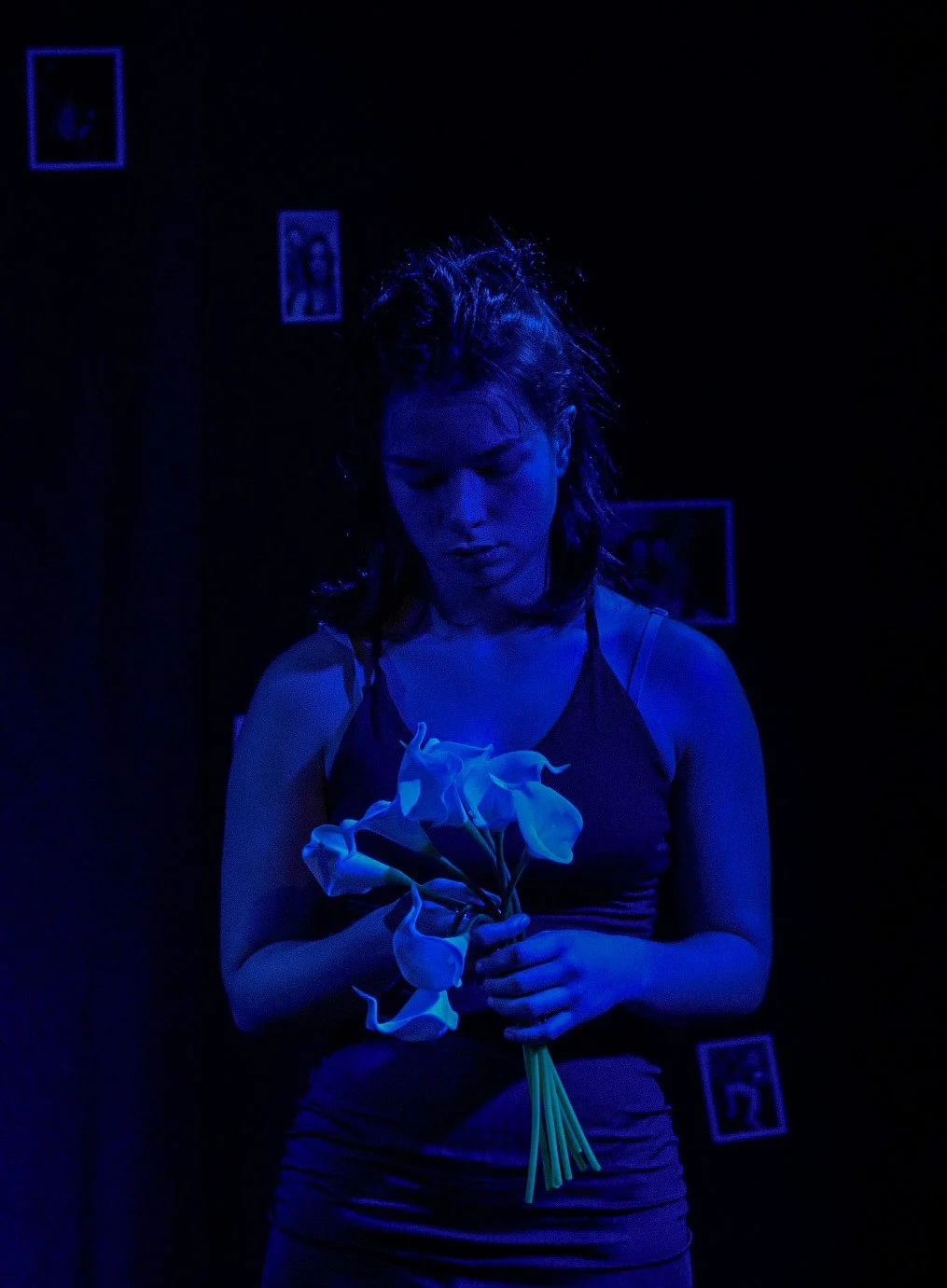 Une femme tient un bouquet de fleurs blanches dans une ambiance sombre avec un éclairage bleu. Des photographies encadrées sont visibles en arrière-plan.