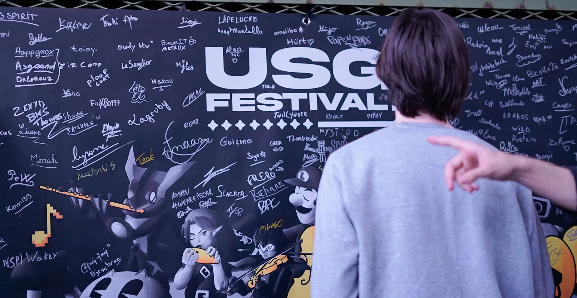 Personne vue de dos regardant un panneau noir avec signatures et dessins, affichant le texte 'USG Festival' en grand, avec divers personnages de jeux vidéo et dessins animés.