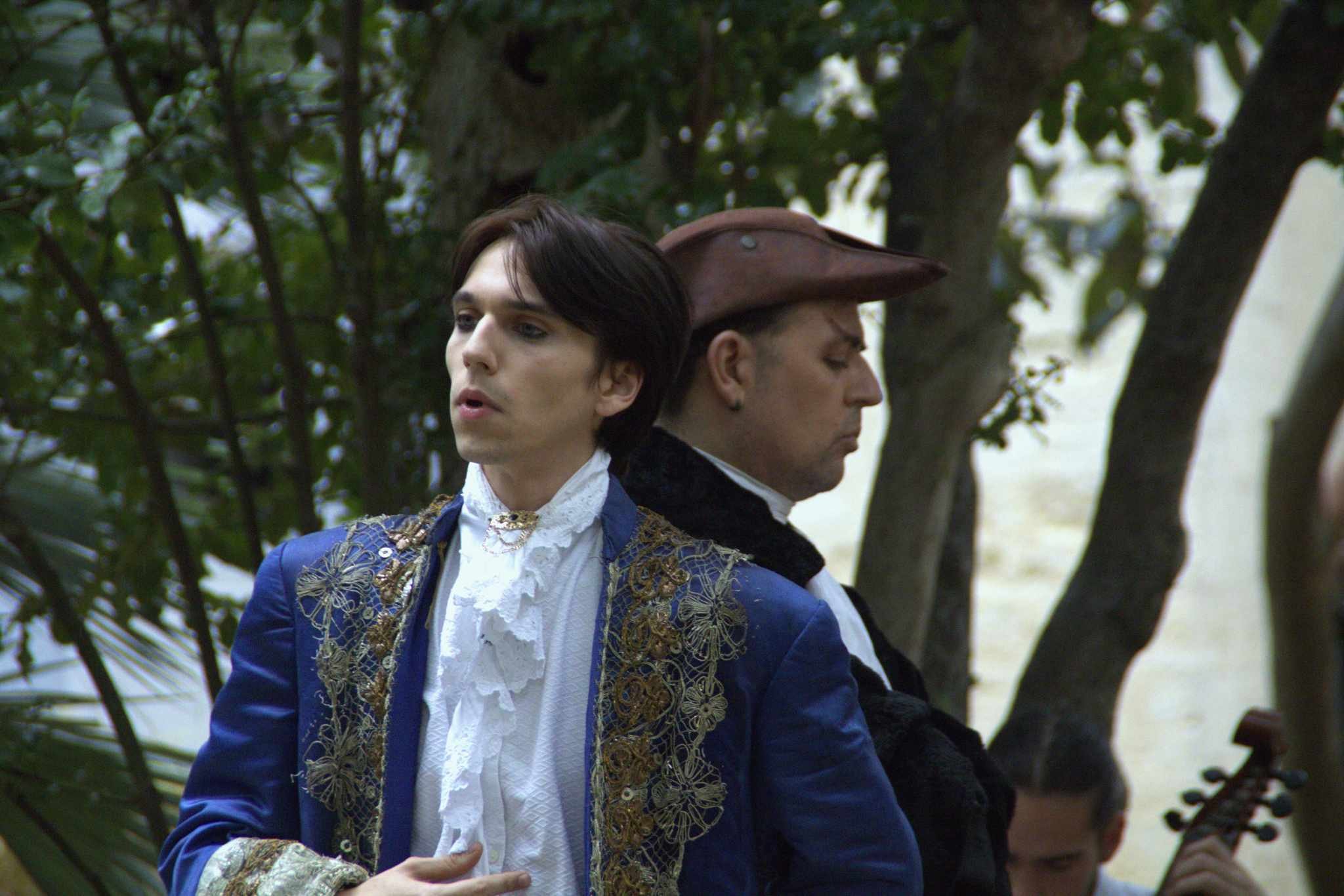 Deux hommes habillés en costumes du XVIIIe siècle, l'un portant un costume bleu avec des broderies dorées et une chemise en dentelle blanche, l'autre portant un chapeau en cuir, dans un décor naturel avec des arbres.