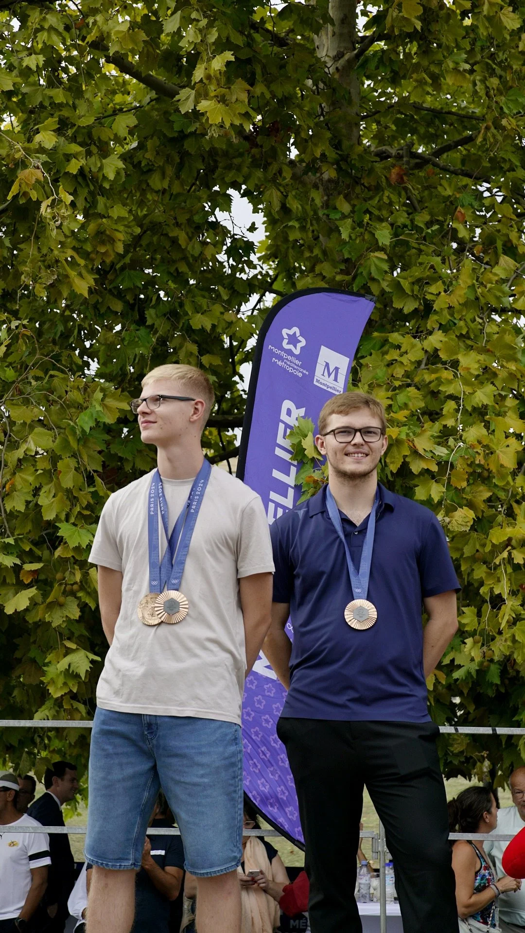 Deux jeunes hommes portant des médailles posent à un podium lors d'une cérémonie de remise de médailles, avec un arbre en arrière-plan.