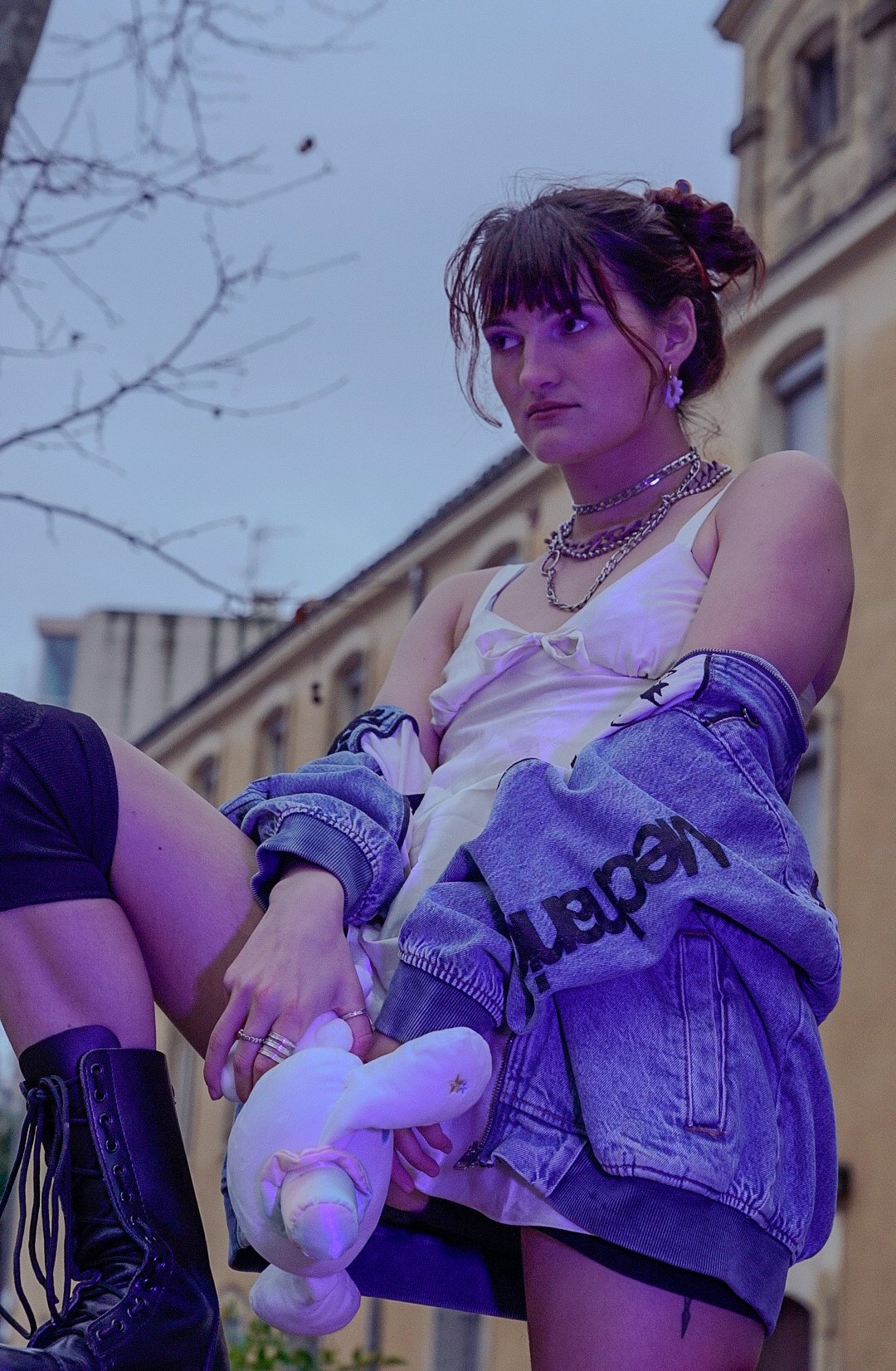 Jeune femme assise avec des vêtements décontractés, portant des bijoux et des chaussettes avec un costume de licorne en peluche, devant un bâtiment urbain lors de soirée ou crépuscule.
