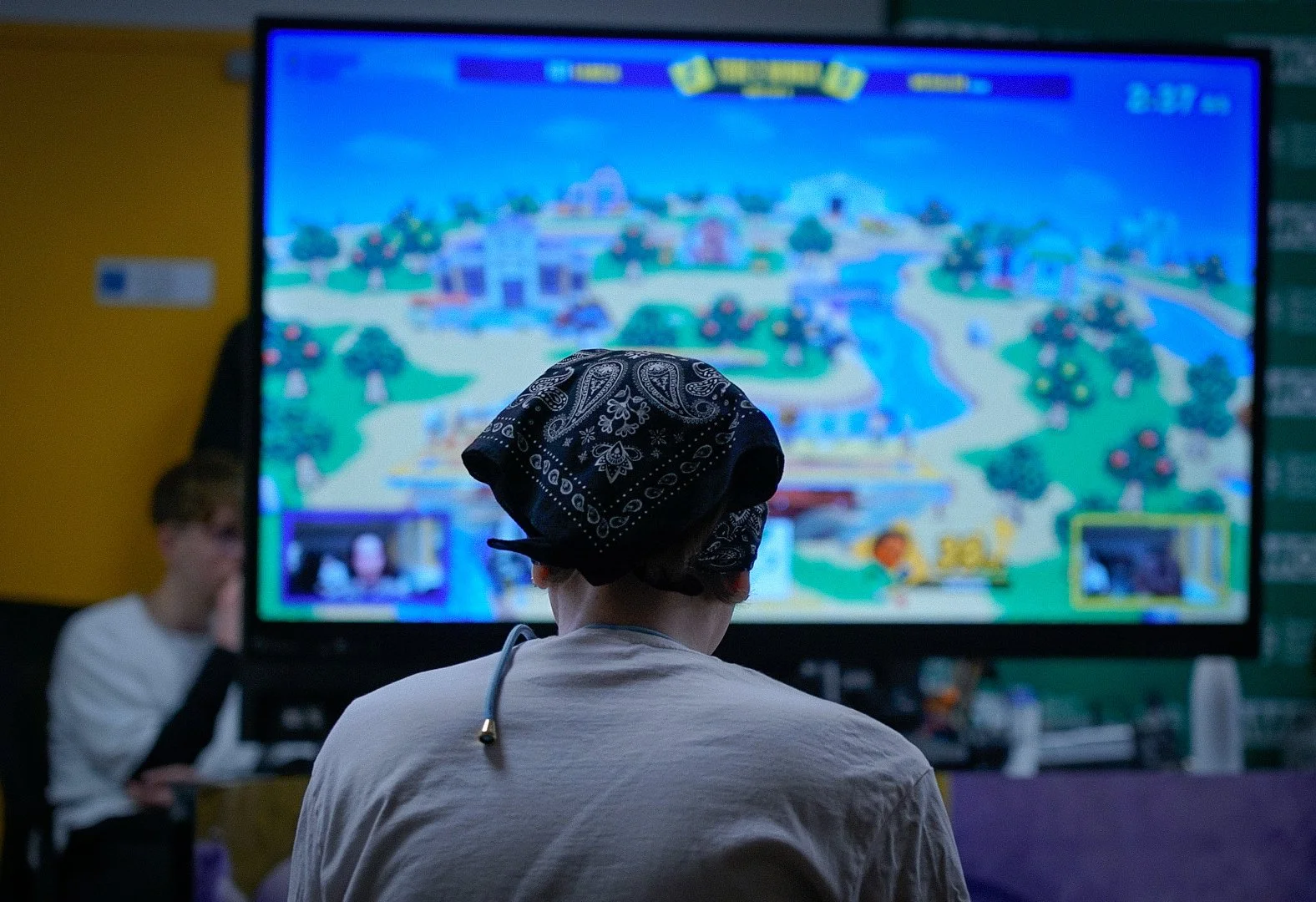 Une personne regarde un grand écran de jeu vidéo, portant un bandana noir avec des motifs blancs, dans une salle sombre.