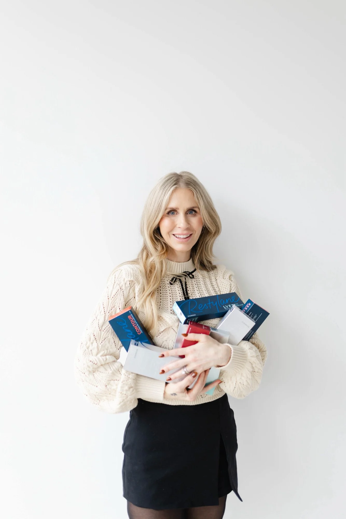 A woman with long blonde hair wearing a cream knit sweater and black skirt, holding several boxes of filler, smiling in front of a plain white wall.