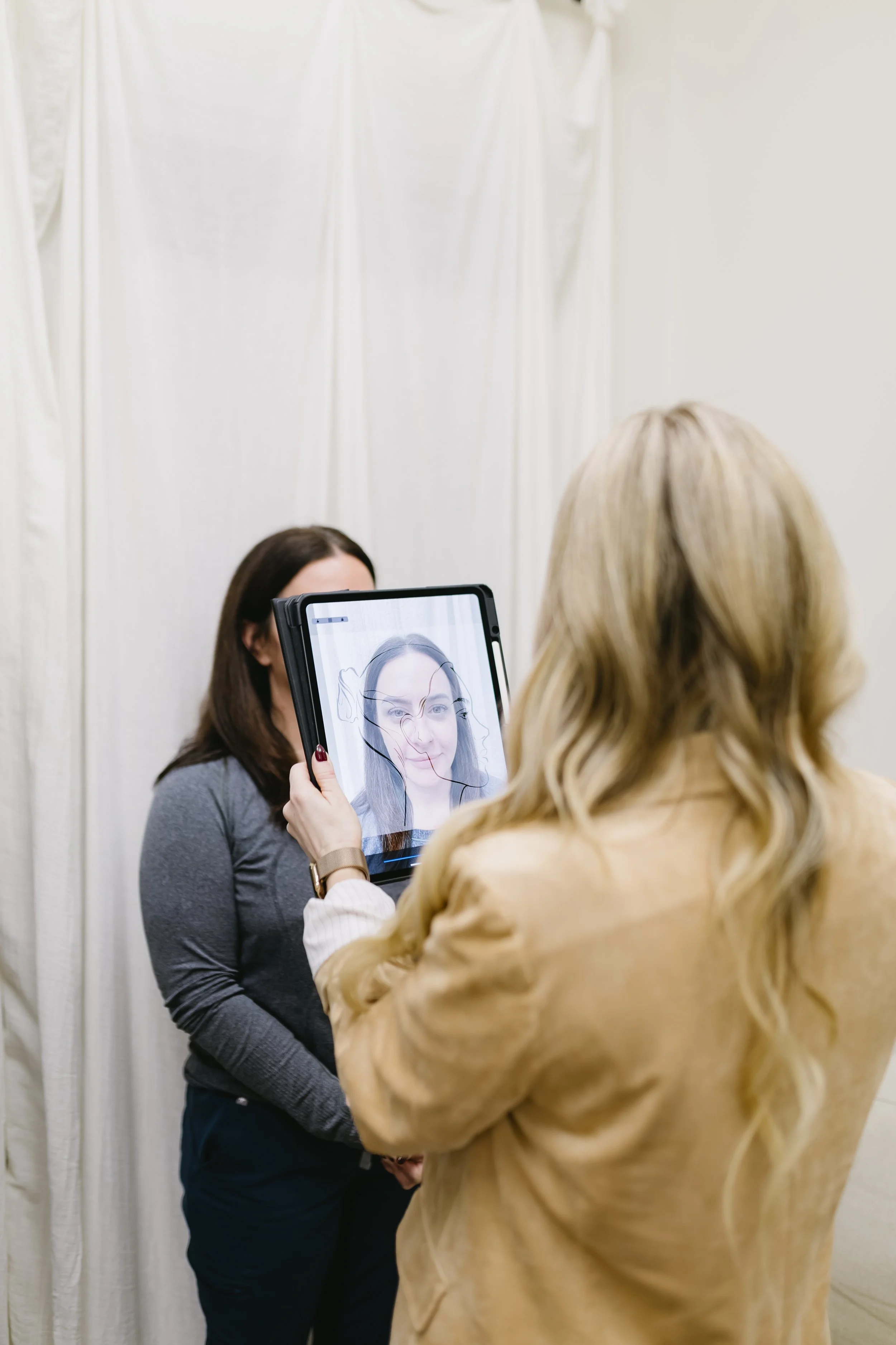 Two women standing in front of a white curtain, one taking a photo of the other with a tablet. The woman with dark hair is facing the camera, and the woman with blonde hair is holding the tablet. Personalized care.