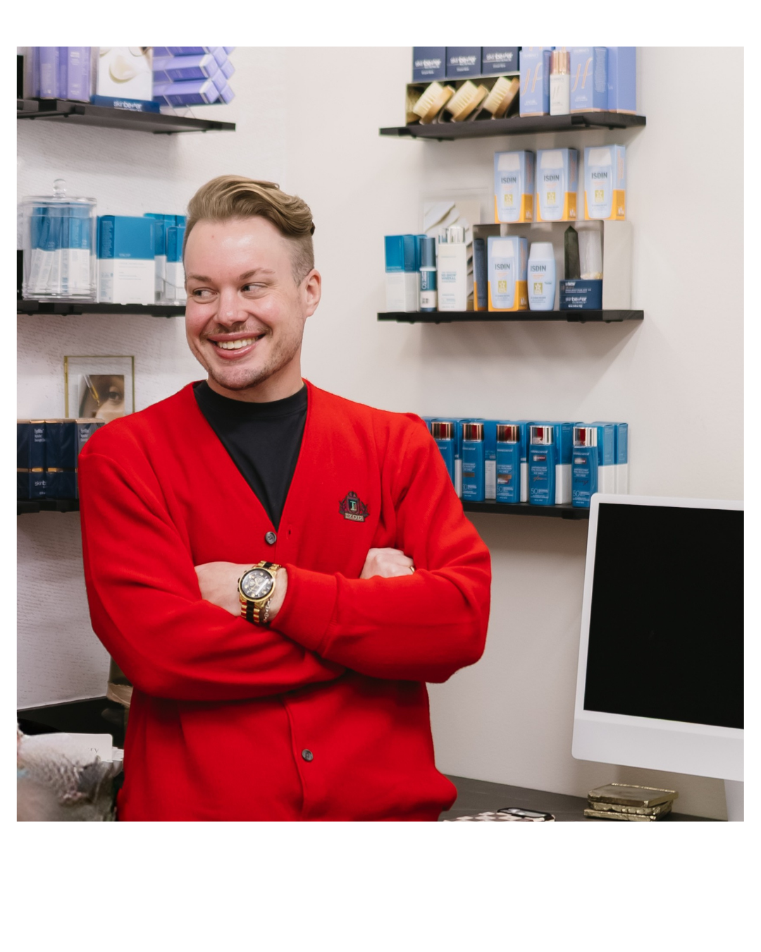 A smiling man with short, styled hair wearing a red cardigan and a black shirt stands with arms crossed in a store with shelves of skincare products behind him.