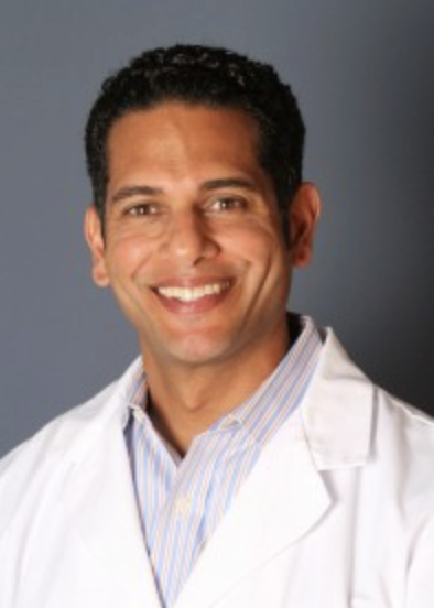 A smiling man in a white medical coat and striped shirt against a gray background.