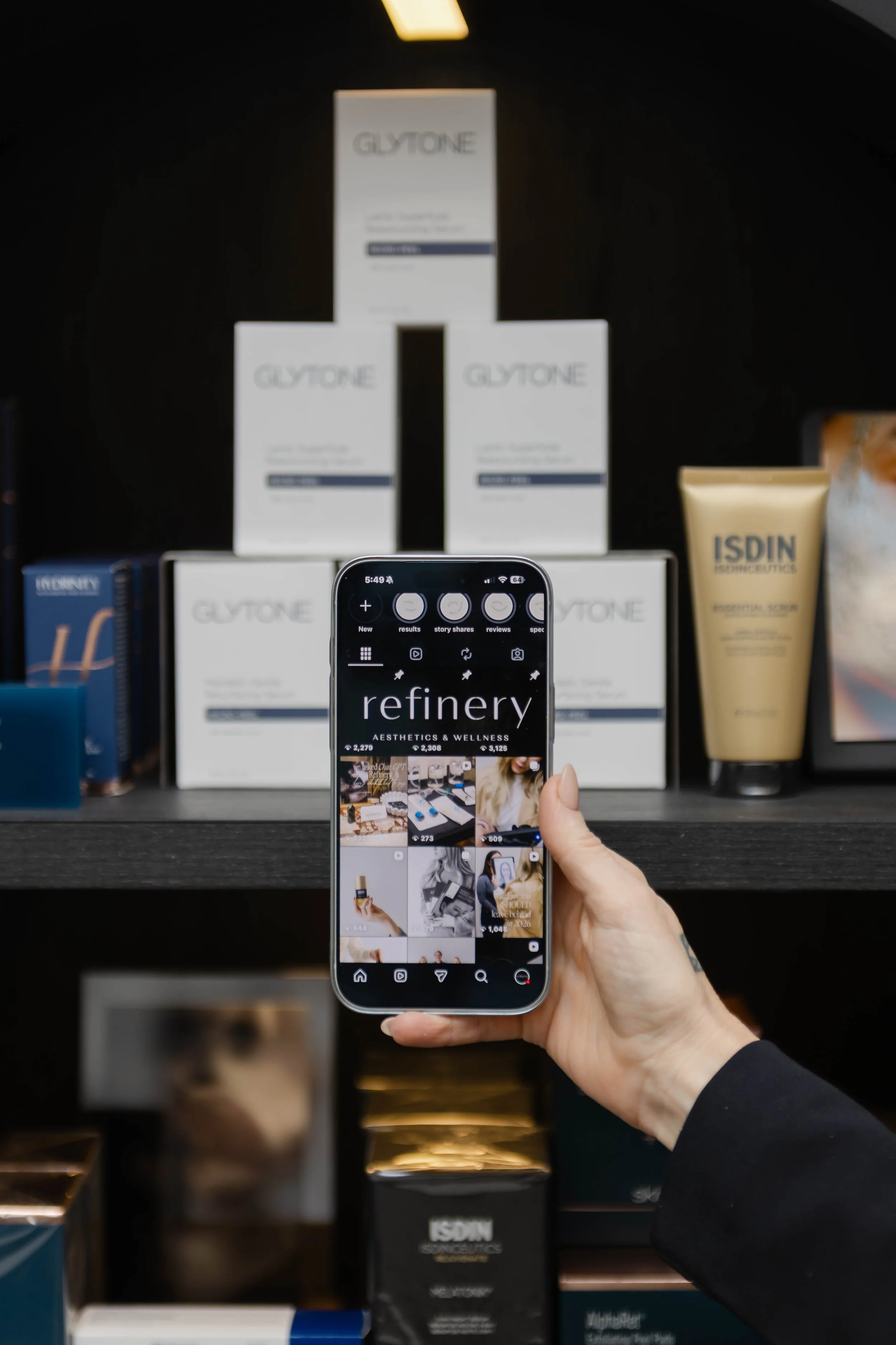 Person holding smartphone taking a photo of various skincare and wellness products displayed on a shelf, including brands Glytone , Skinbetter Science, Hydrinity, Colorscience, Obagi Medical and ISDIN.