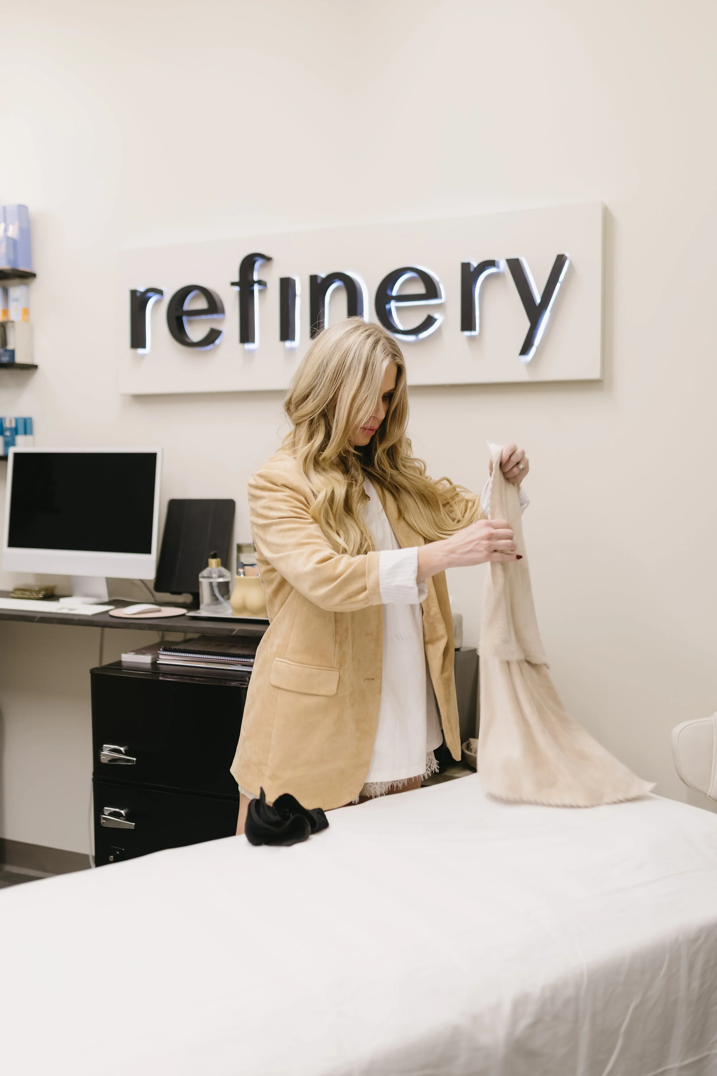 A woman with long blonde hair, wearing a tan blazer, is examining a beige scarf or clothing item in a retail store with a "refinery" sign on the wall. She is setting the bed up for a dermaplane and facial.