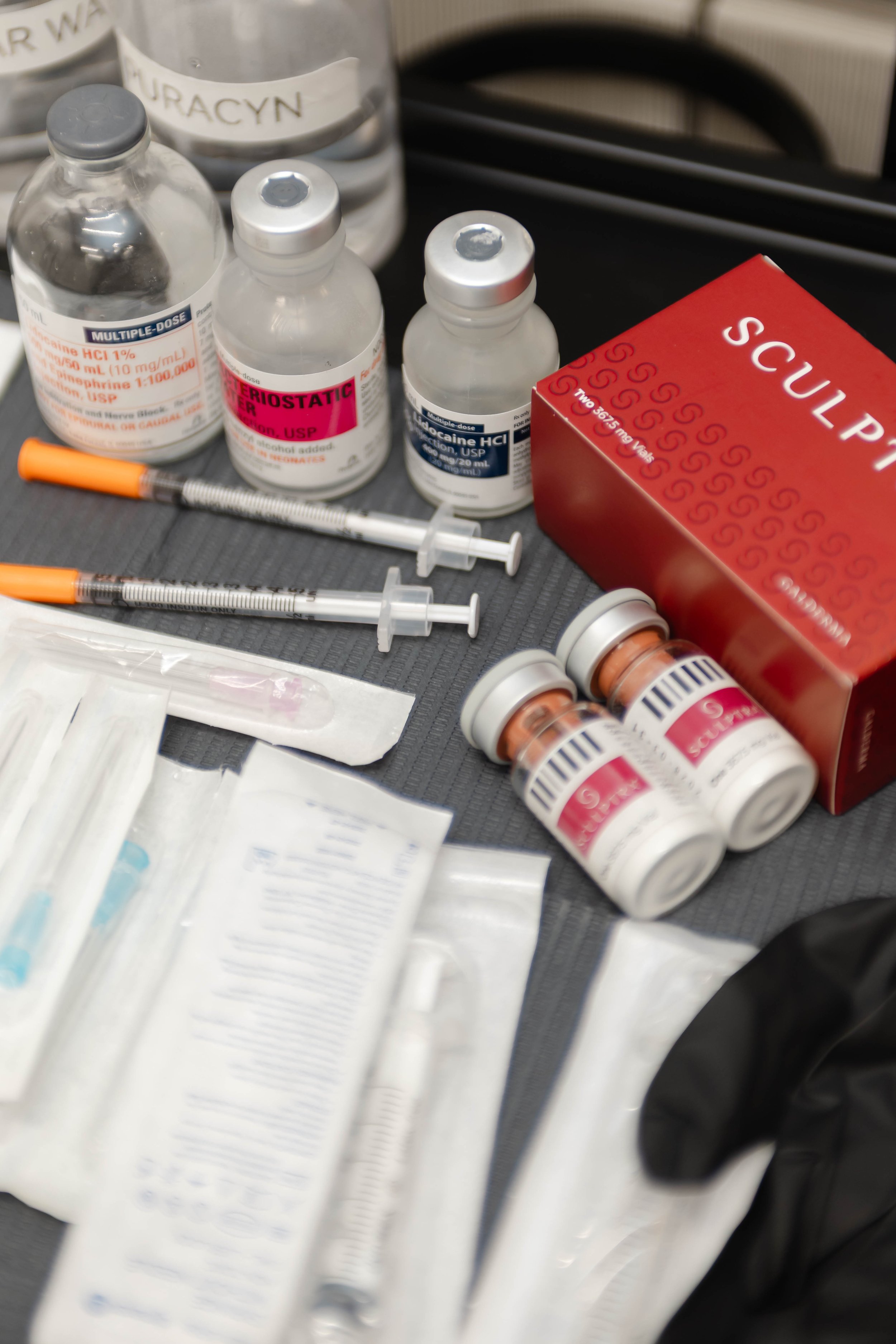 Medical supplies including syringes, vials, bottles of medication, and a box of surgical masks on a black surface. There is Sculptra and Dysport on the try.
