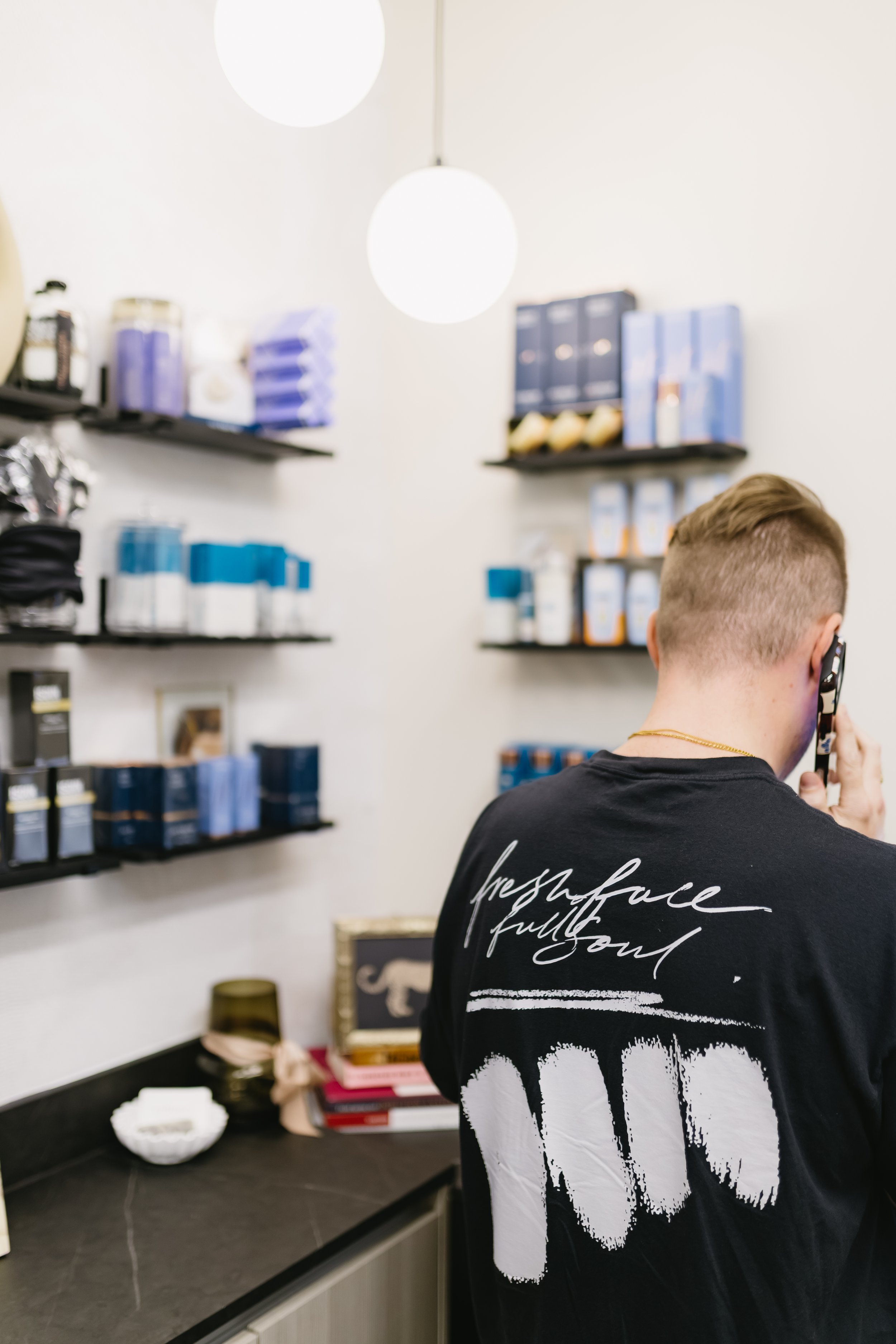 A person talking on the phone at Refinery Aesthetics & Wellness. His shirt says fresh face full soul. The shelves have Isdin, Hydrinity, Colorscience and Skinbetter products.