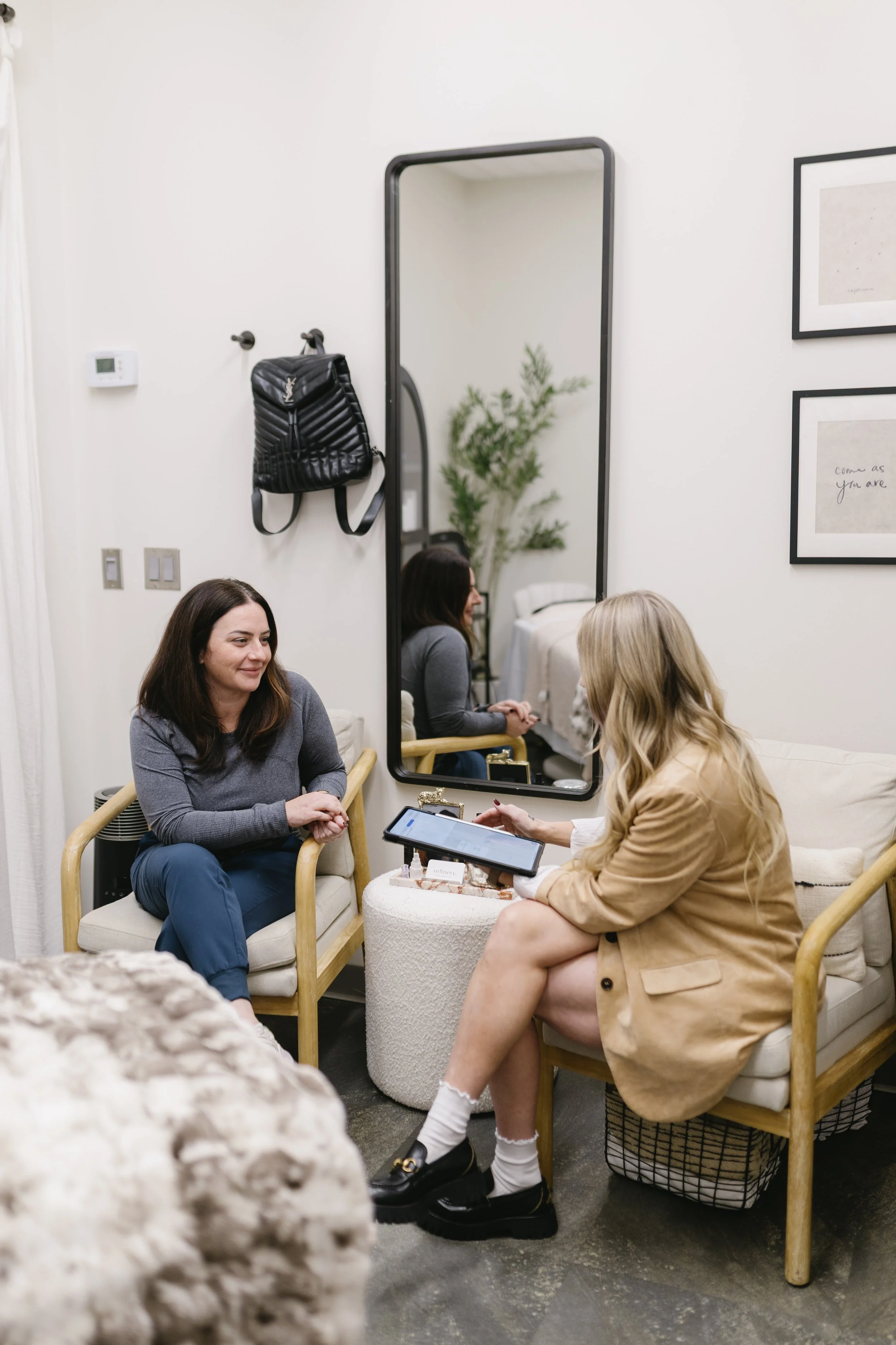 Two women sitting and talking in a cozy, modern room with a large mirror on the wall, a framed art piece, and a small table with a tablet during a skin rejuvenation consultation.