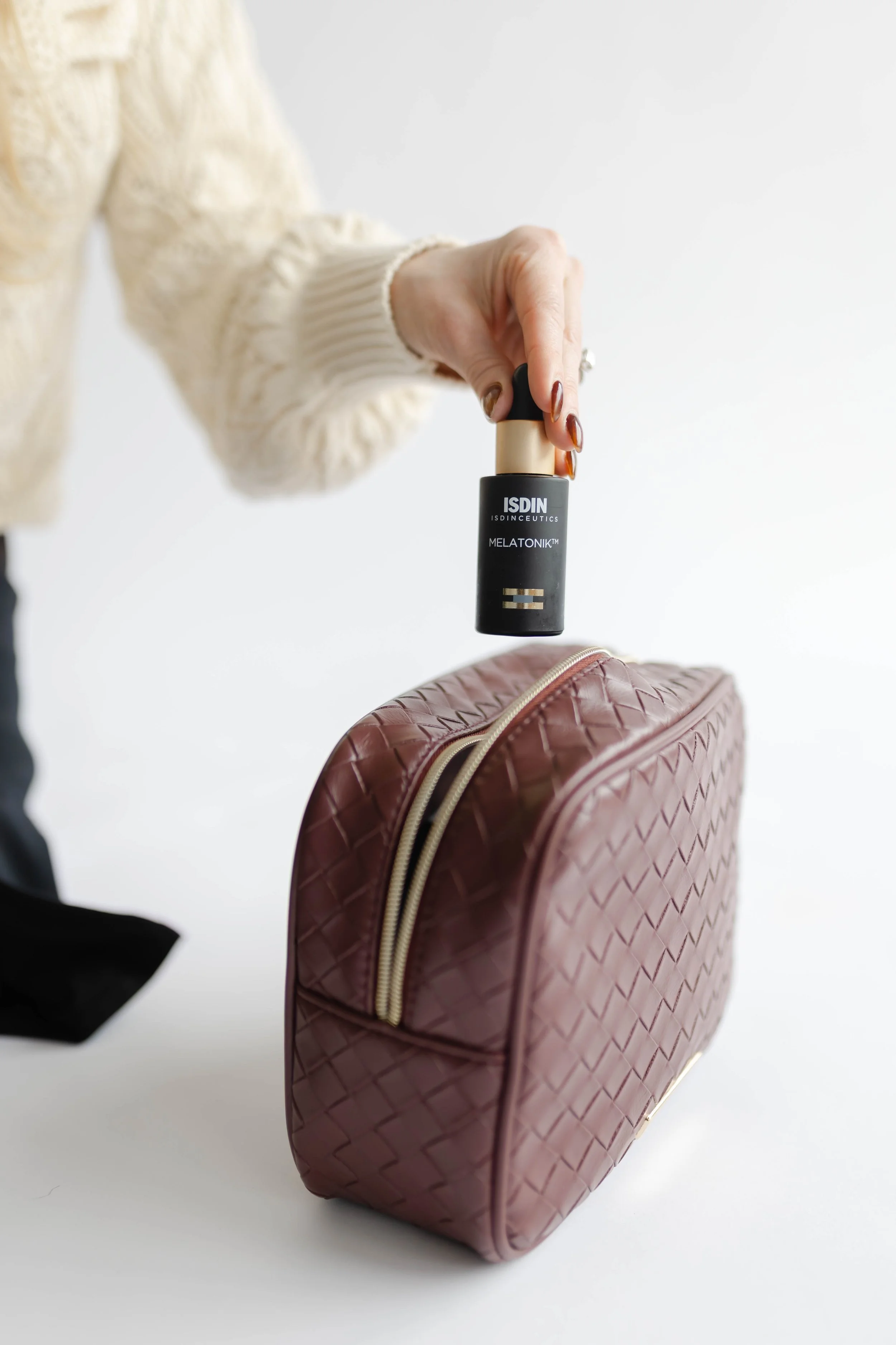 A person in a cream-colored knit sweater is holding a bottle of ISDIN Melatonin sleeping spray above a burgundy quilted bag with gold zippers on a white background.