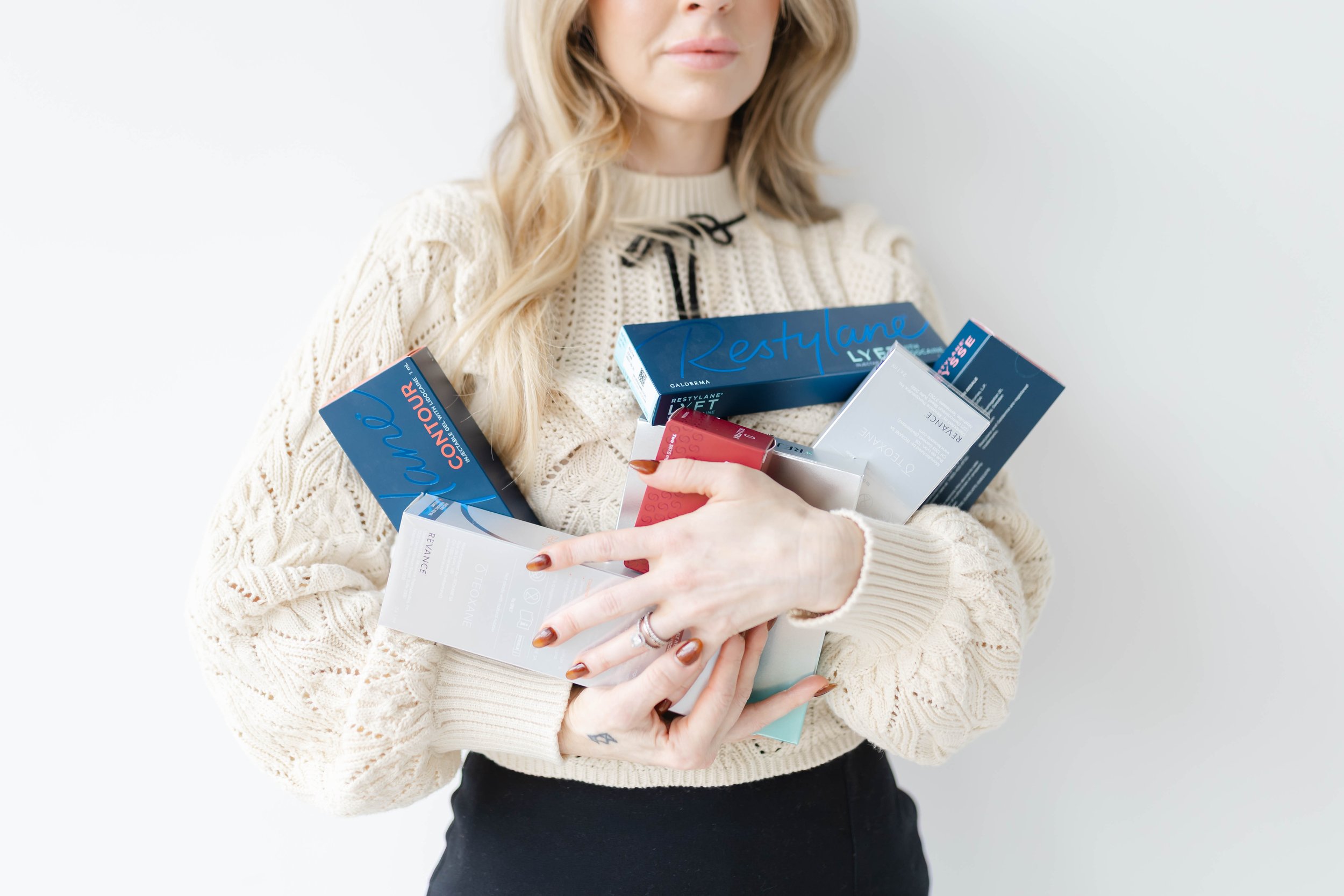 Holly Tabor, MS, APRN, FNP-C, a woman wearing a cream-colored knitted sweater, holding multiple skincare product boxes, including Restylane, RHA, Dysport, Jeuveau, Daxxify, Rejuran, exosomes, against a plain light background.