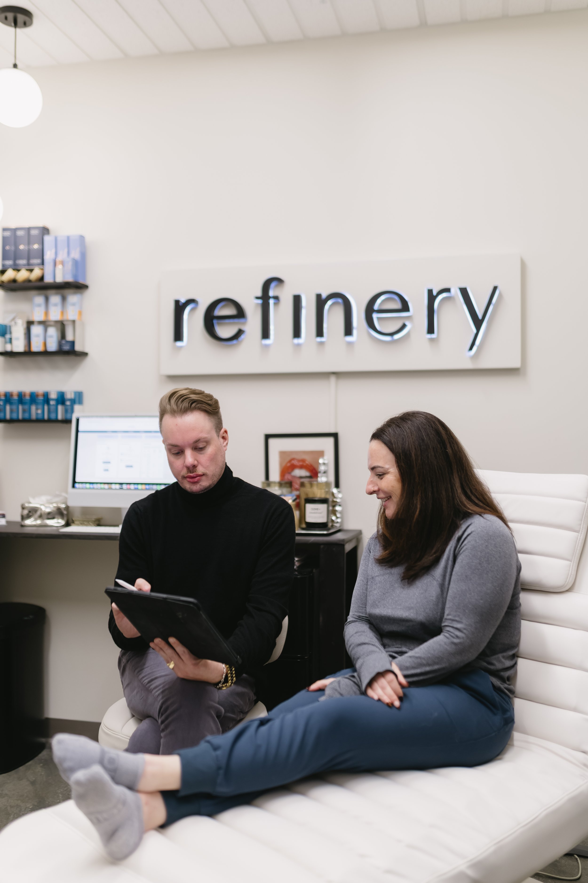 Two people sitting on a white leather couch in a modern office, looking at a tablet together, with a sign that says 'refinery' on the wall behind them. The male is showing the patient Skinbetter Alto Advanced and discussing skincare.