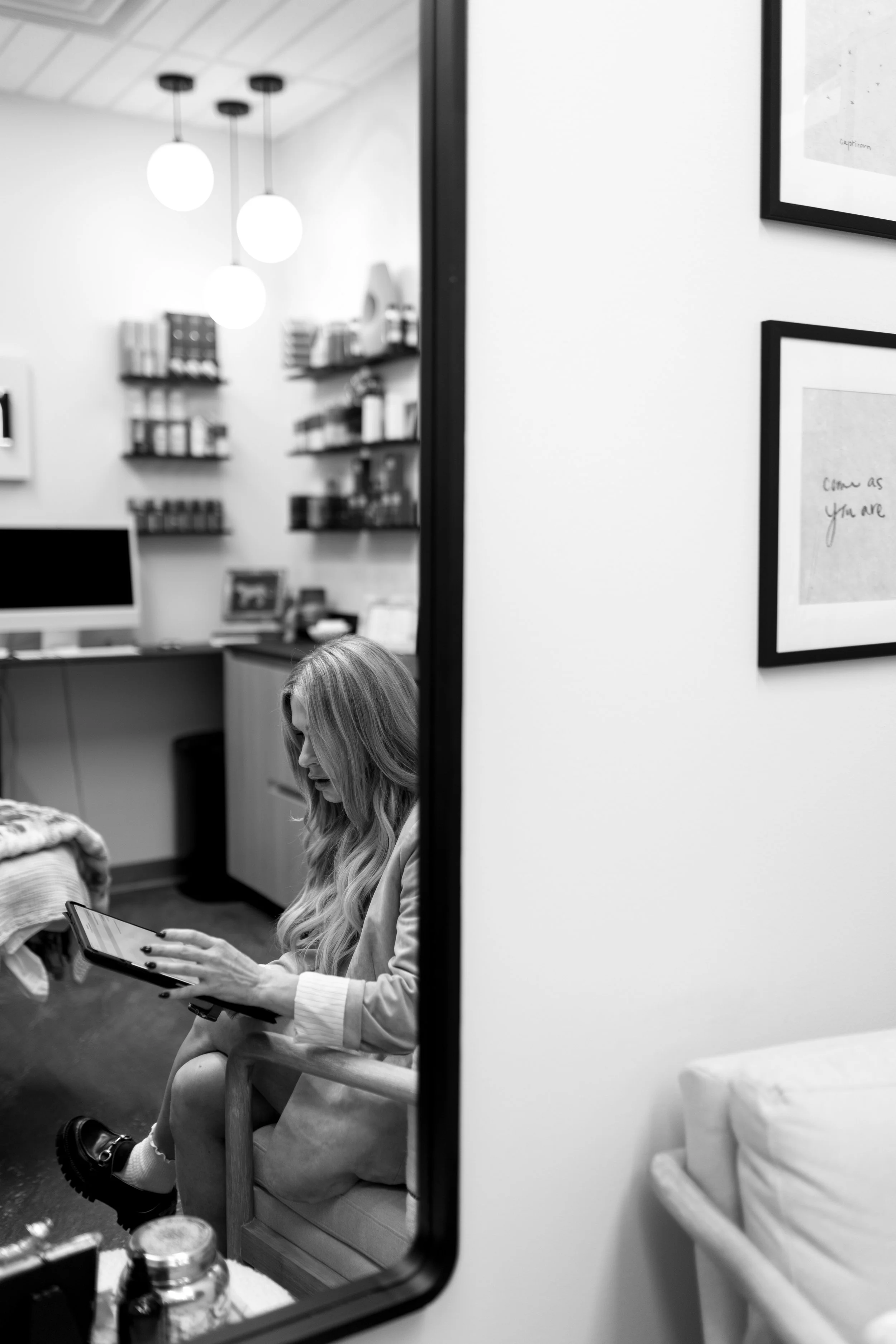 A woman with long wavy hair sitting in a chair, looking at a tablet, reflected in a mirror at Refinery Aesthetics & Wellness, shelves with books and documents, and framed pictures on the wall.