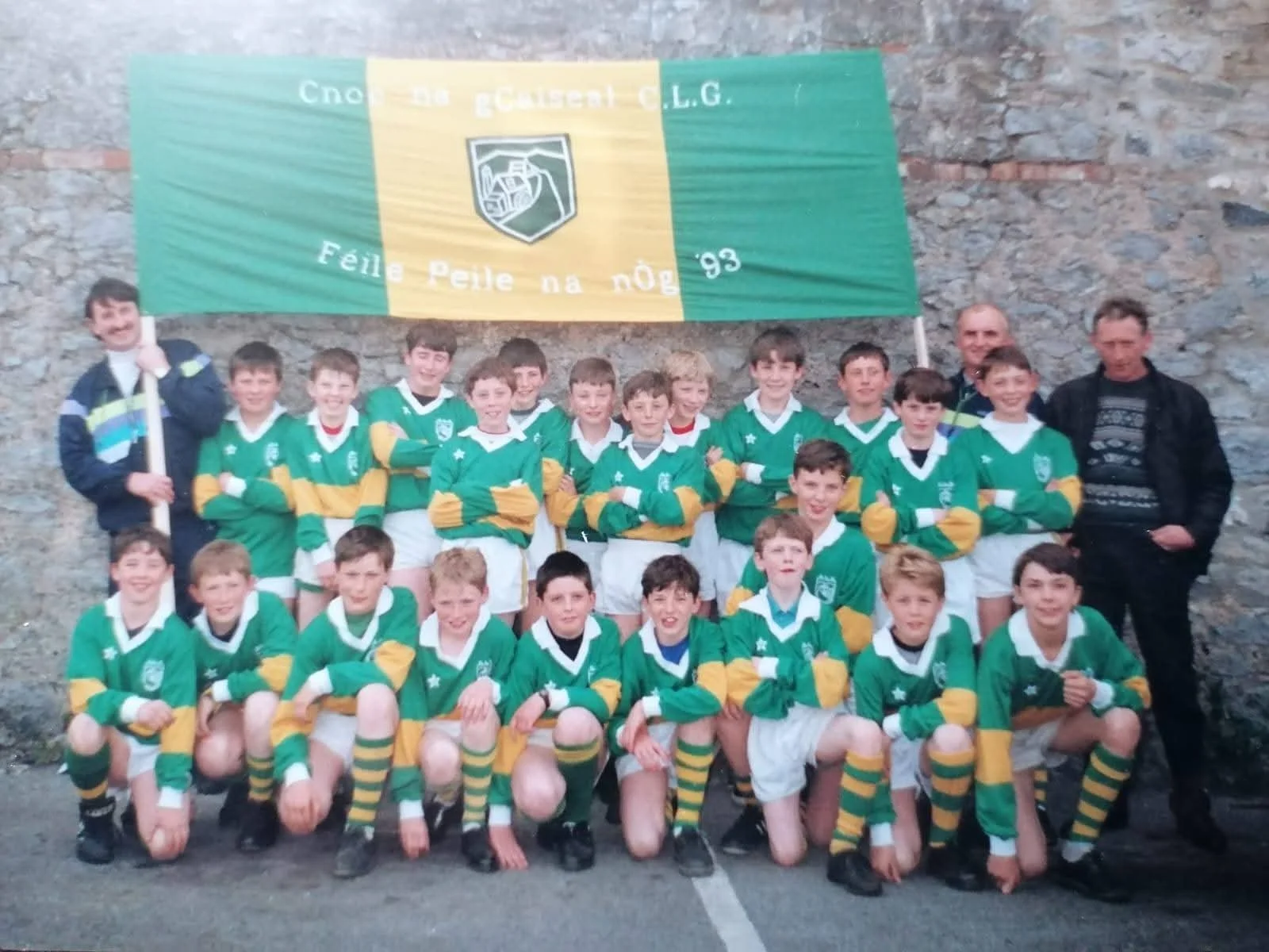 A group of young boys in green and yellow rugby uniforms posing for a team photo with two men wearing jackets, standing in front of a stone wall and holding a large team banner that has a crest with a rugby ball and the words 'Feile Peile na nOg'.