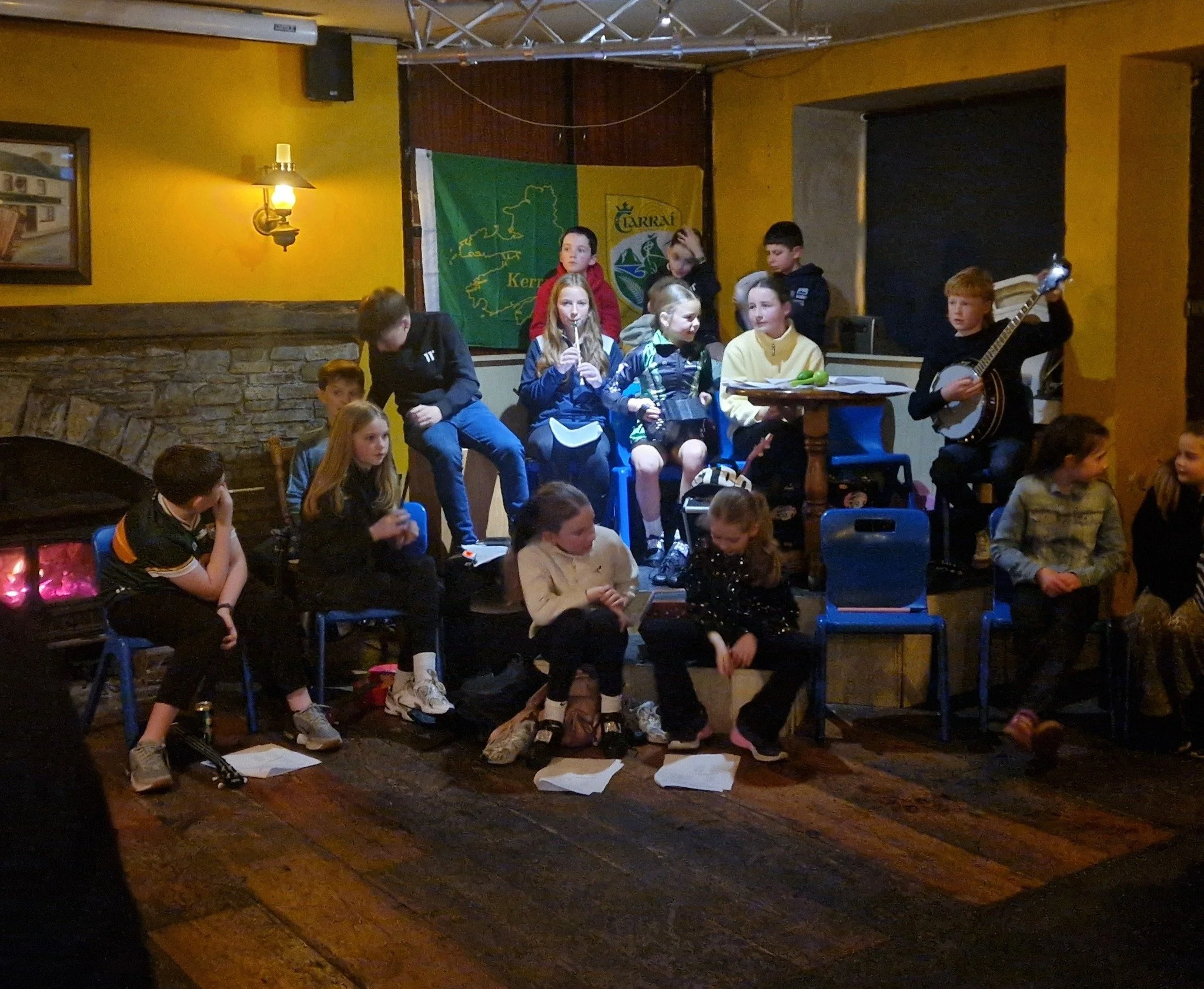 A group of children sitting and standing on a wooden stage in a cozy, dimly lit restaurant or pub. Some children are holding paper and a guitar, indicating they may be preparing for or participating in a performance. The background features yellow walls, decorative banners, and a stone accent wall.