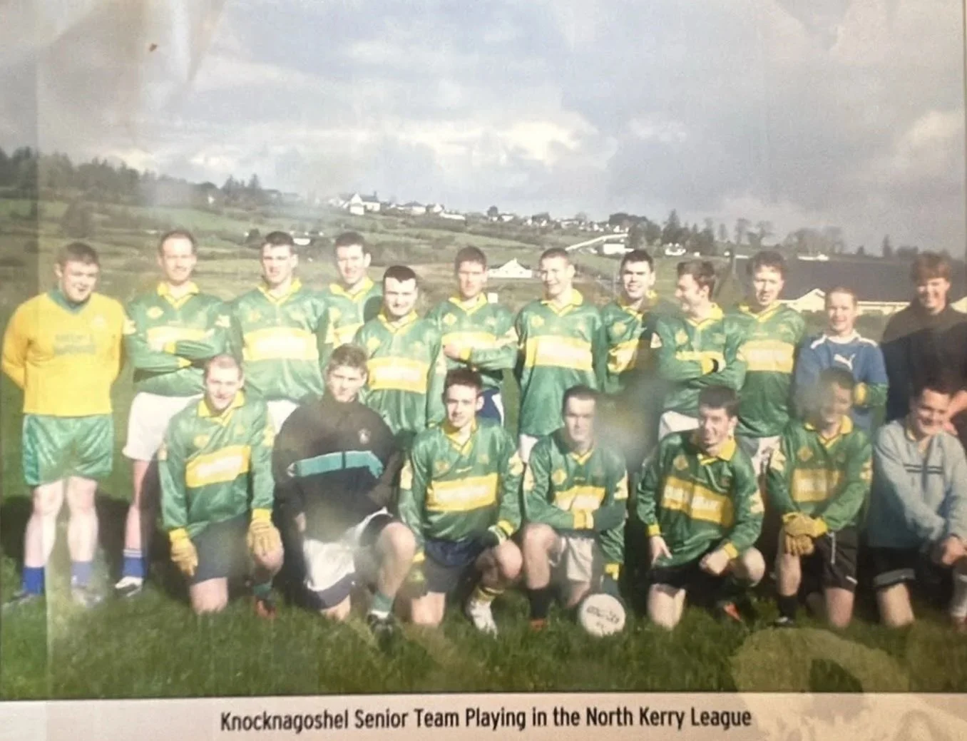 A group of young men in green and yellow sports uniforms posing outdoors for a team photo, resembling a Gaelic football team, with some kneeling in the front row and others standing in the back, against a backdrop of rolling hills and houses.