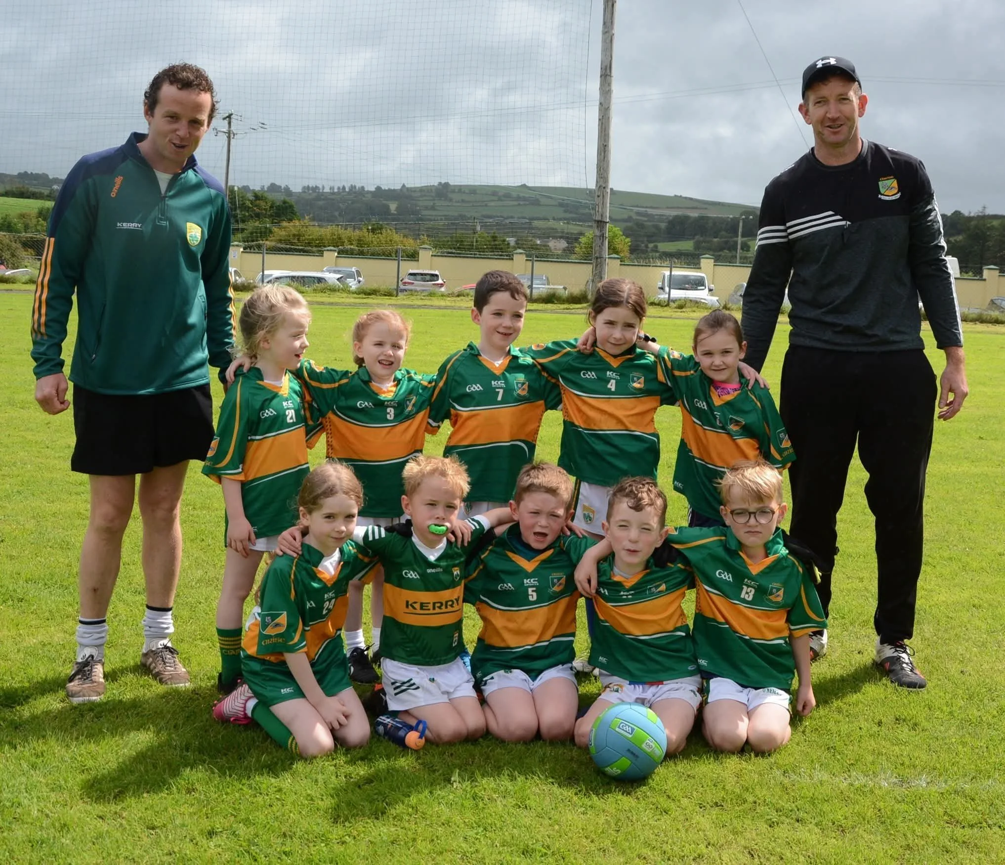 Group of young children and two adult coaches posing for a team photo on a grassy field during daytime, with hills, a fence, and parked cars in the background.