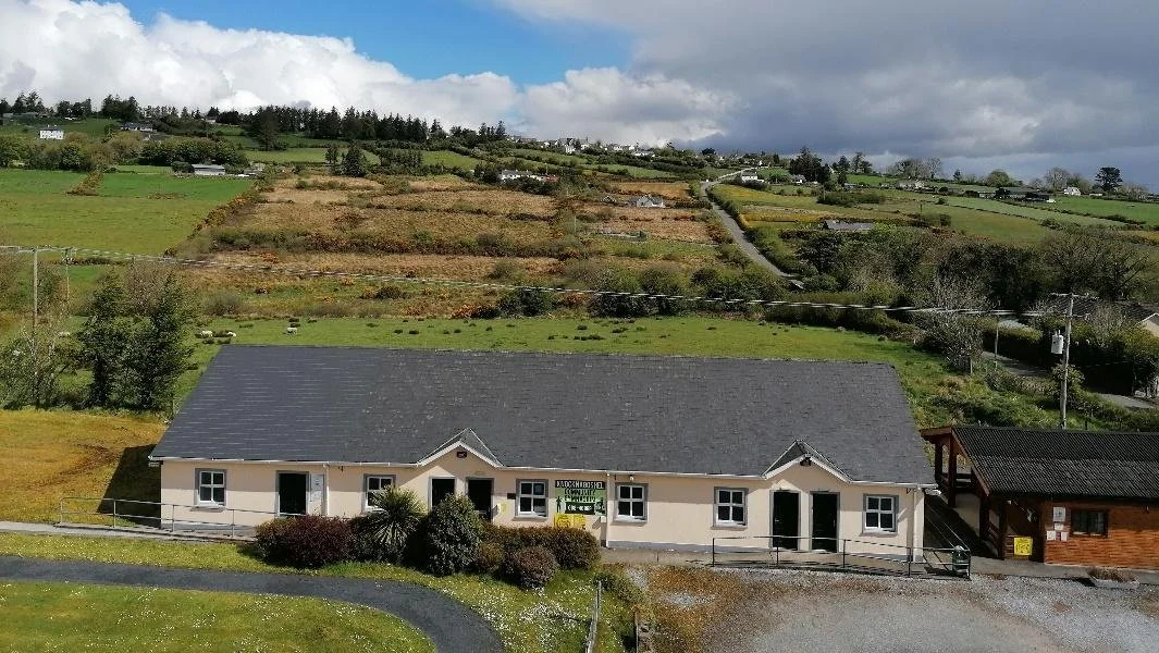 A single-story house with a dark gray roof and light-colored walls, situated in a rural area with rolling hills covered in greenery and patches of farmland in the background.