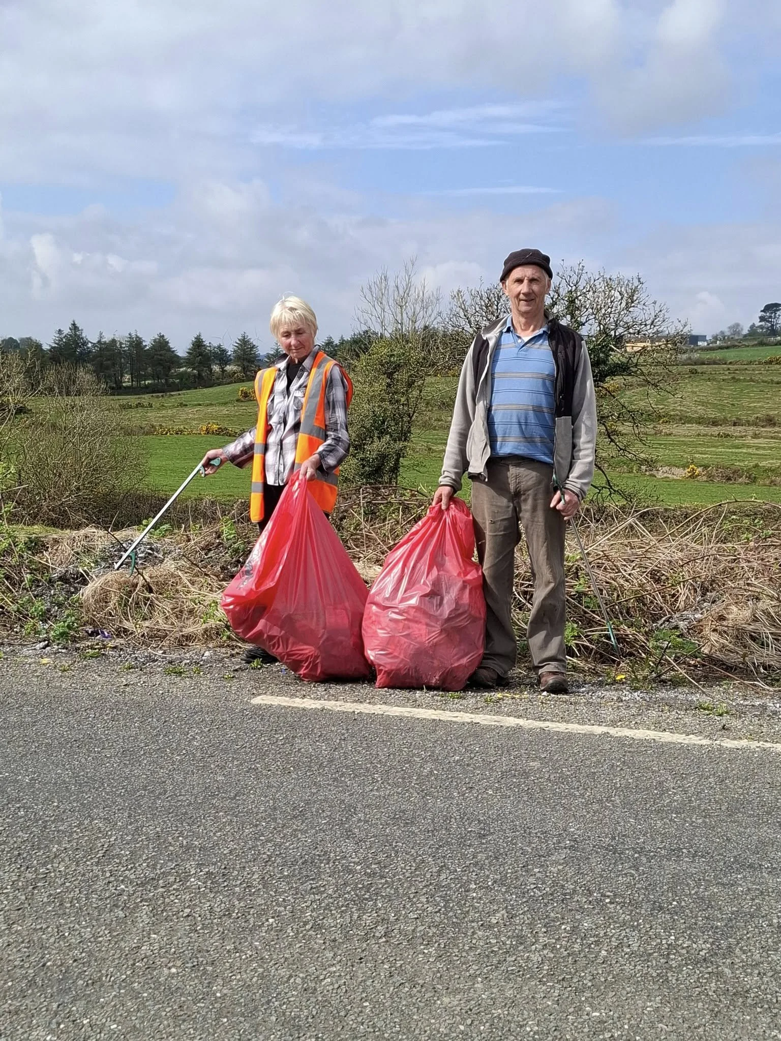 Two elderly people, a woman on the left with white hair wearing a high-visibility vest and holding trash pick-up tools, and a man on the right in a hat and outdoor clothing, standing beside large red garbage bags filled with collected litter on a roa