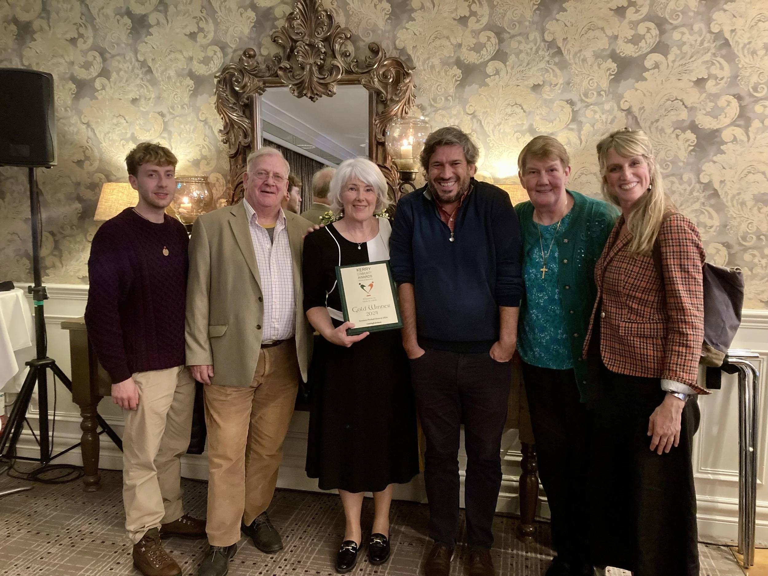 A group of six people standing together at an indoor event, with a decorative mirror and ornate wallpaper in the background. One person is holding a framed award or certificate.