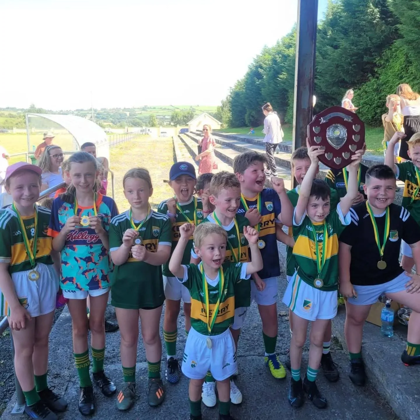 Group of young children in sports uniforms celebrating with medals and a shield-shaped trophy after a sports game or event outdoors.