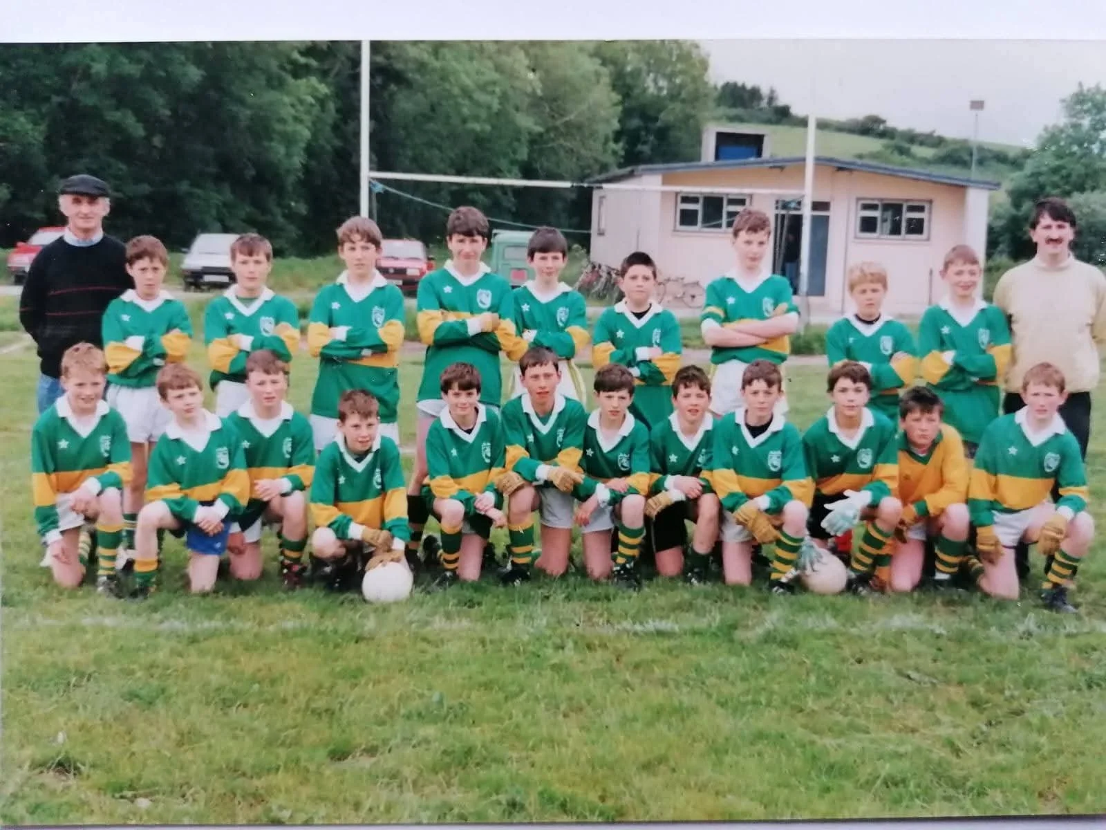 A youth rugby team in green and yellow uniforms, with two adult coaches, posing on a grassy field with a rugby ball in front. There is a small building and cars in the background.