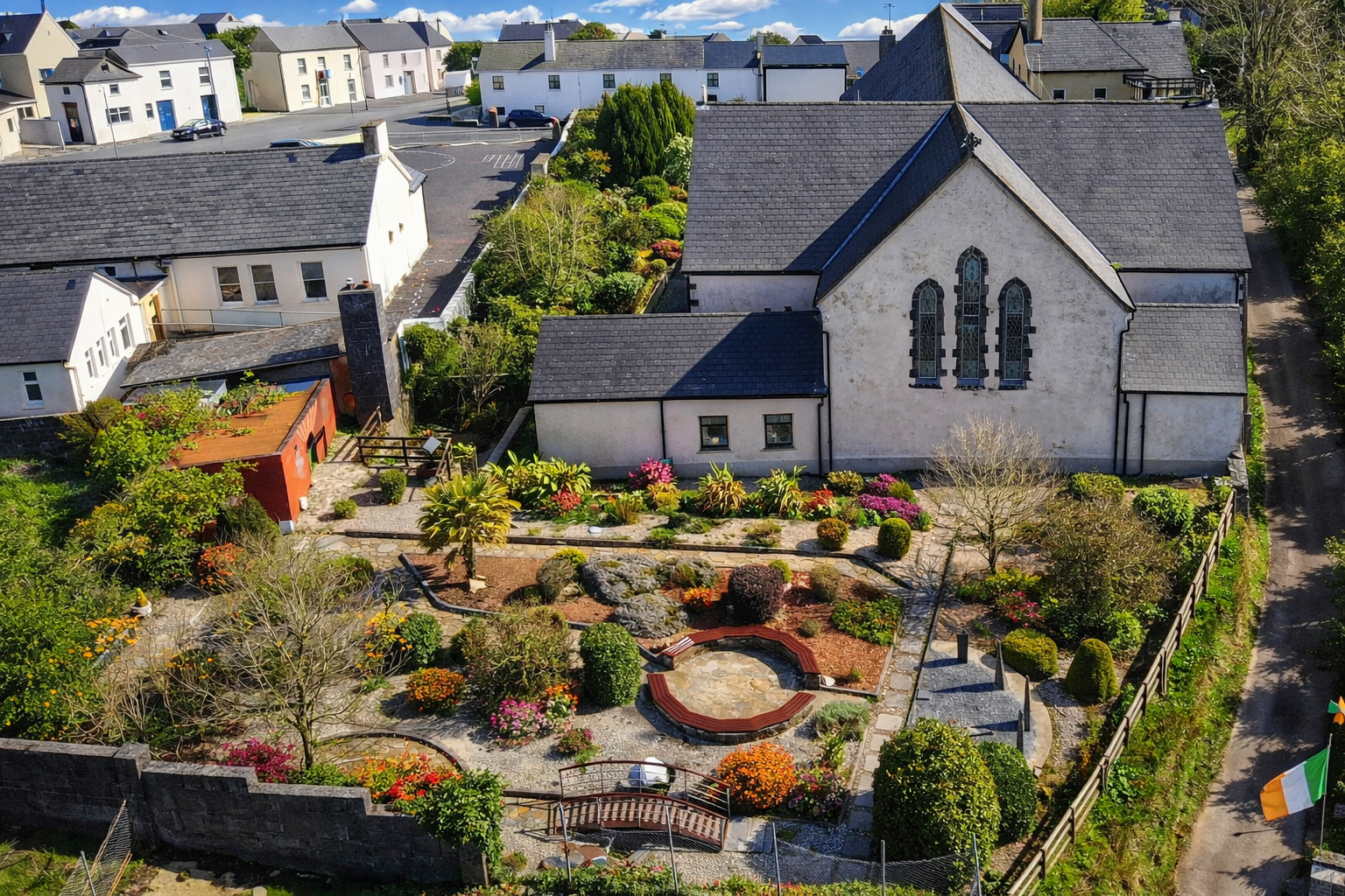 Bird's eye view of a church with a small garden in front, featuring colorful flowers, small trees, bushes, and a pond, with neighboring houses and a parking lot in the background.