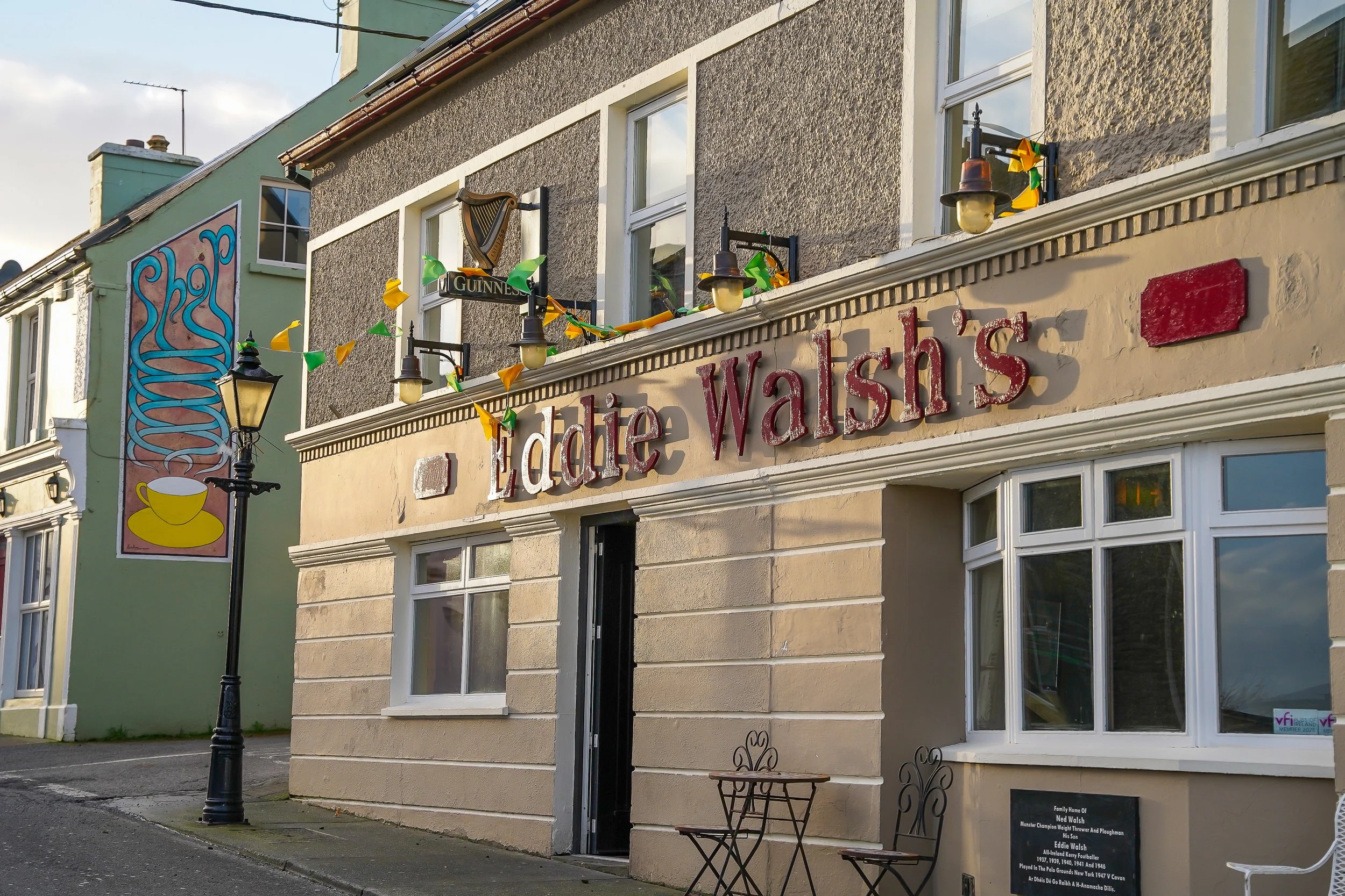 Street view of a cafe with a sign reading 'Eatdie Walsh's' and decorative lighting. There is a mural on an adjacent building showing a harp and a yellow cup. A black street lamp and a small metal table and chairs are in front.